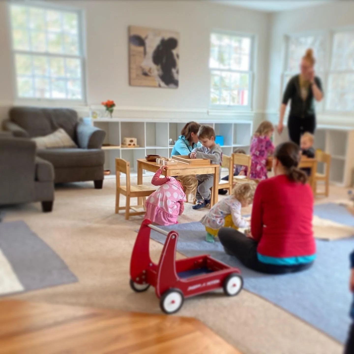 Children playing and adults supervising in a bright indoor playroom with windows, a couch, and shelves.