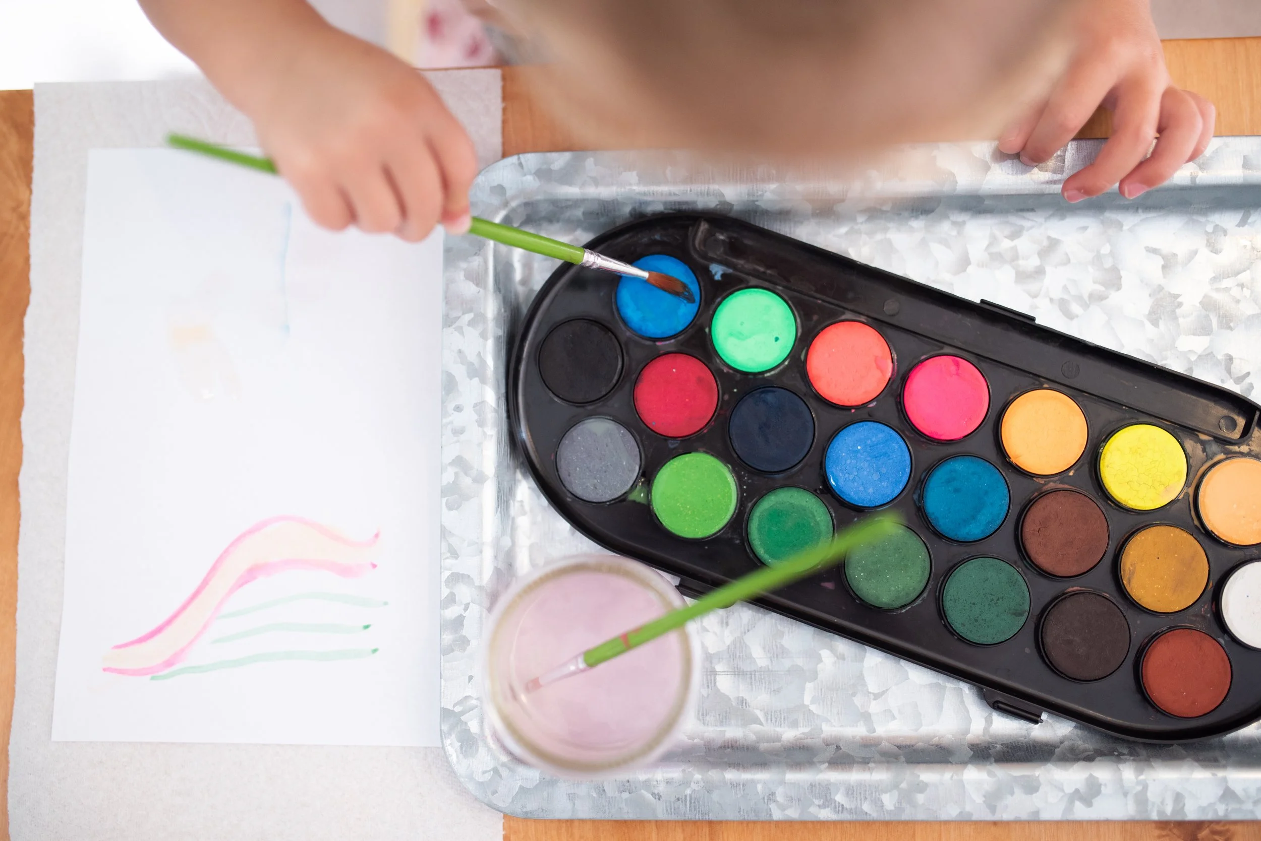 Child painting with a paintbrush in a small cup of water, watercolor paints on a tray, and a piece of paper with colorful streaks.