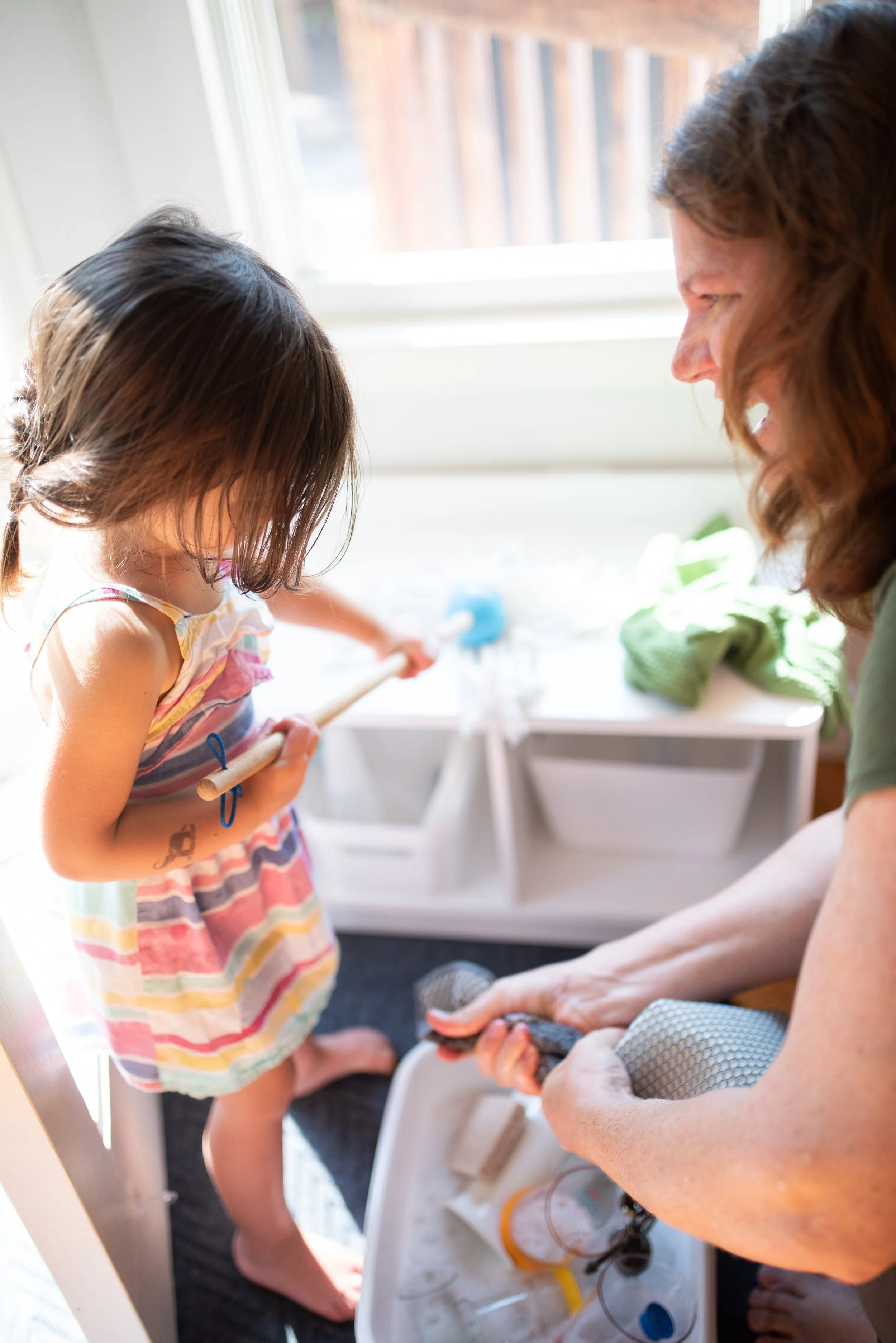 A young girl with brown hair wearing a colorful striped dress and a woman with brown hair working together by a sink, with cleaning supplies and a window in the background.