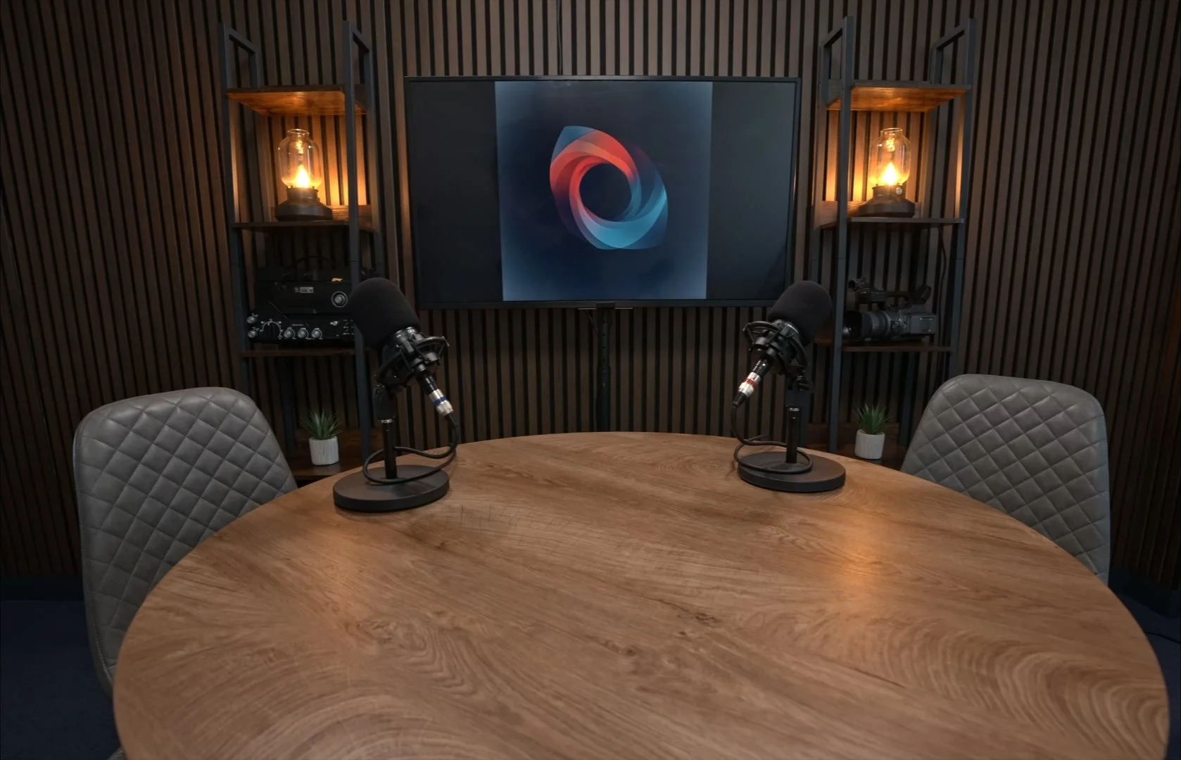 A podcast recording studio with a round wooden table, two microphones, two quilted chairs, a large screen with a colorful abstract logo, and two shelves with lamps, plants, and audio equipment in the background.
