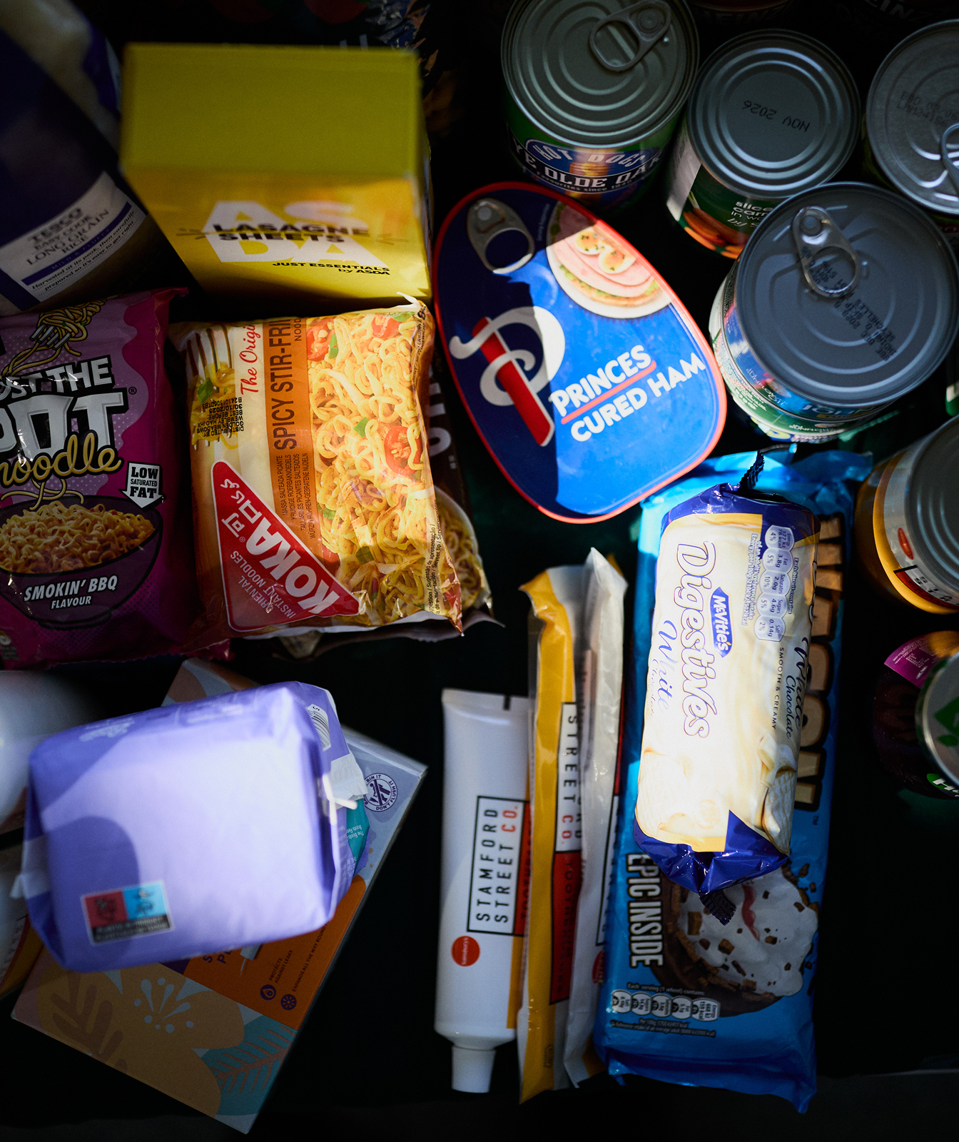 There is a collection of foods - some are in packets, cans, bottles and bags. It includes things like noodles, tuna, biscuits and beans.