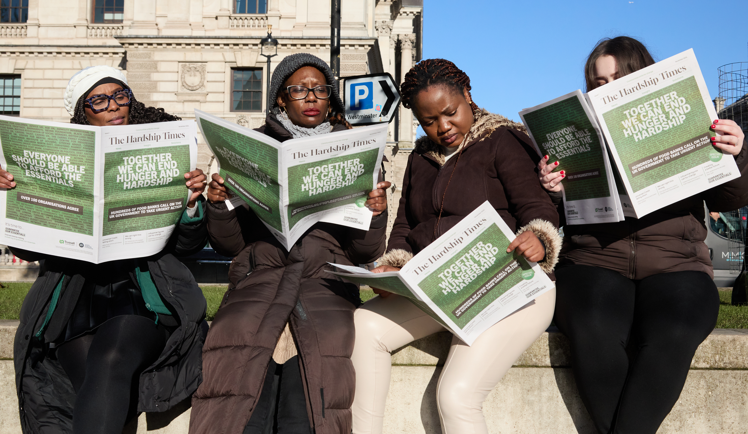 An image of a group of women, sat on a wall. They are all reading the newspaper, which has a green image on the front of it.