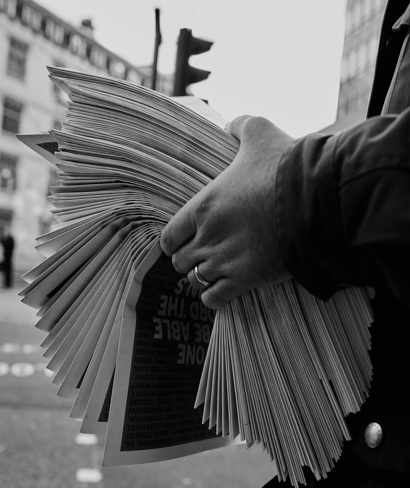 In black and white, someone is holding a stack of newspapers very tightly. You can't see their face - just their hands and the papers. In the background is a street or road crossing.