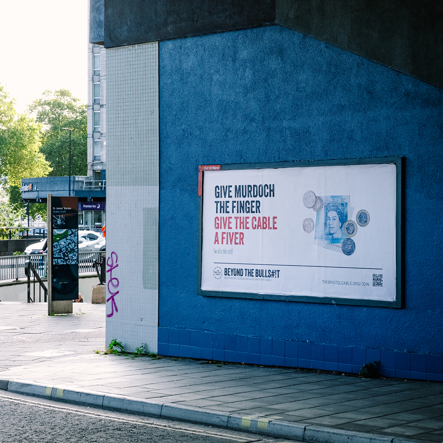 An underpass, with a russian blue wall has a billboard on it. The billboard has an advert for The Bristol Cable which reads 'Give Murdoch the finger, Give the Cable a fiver'