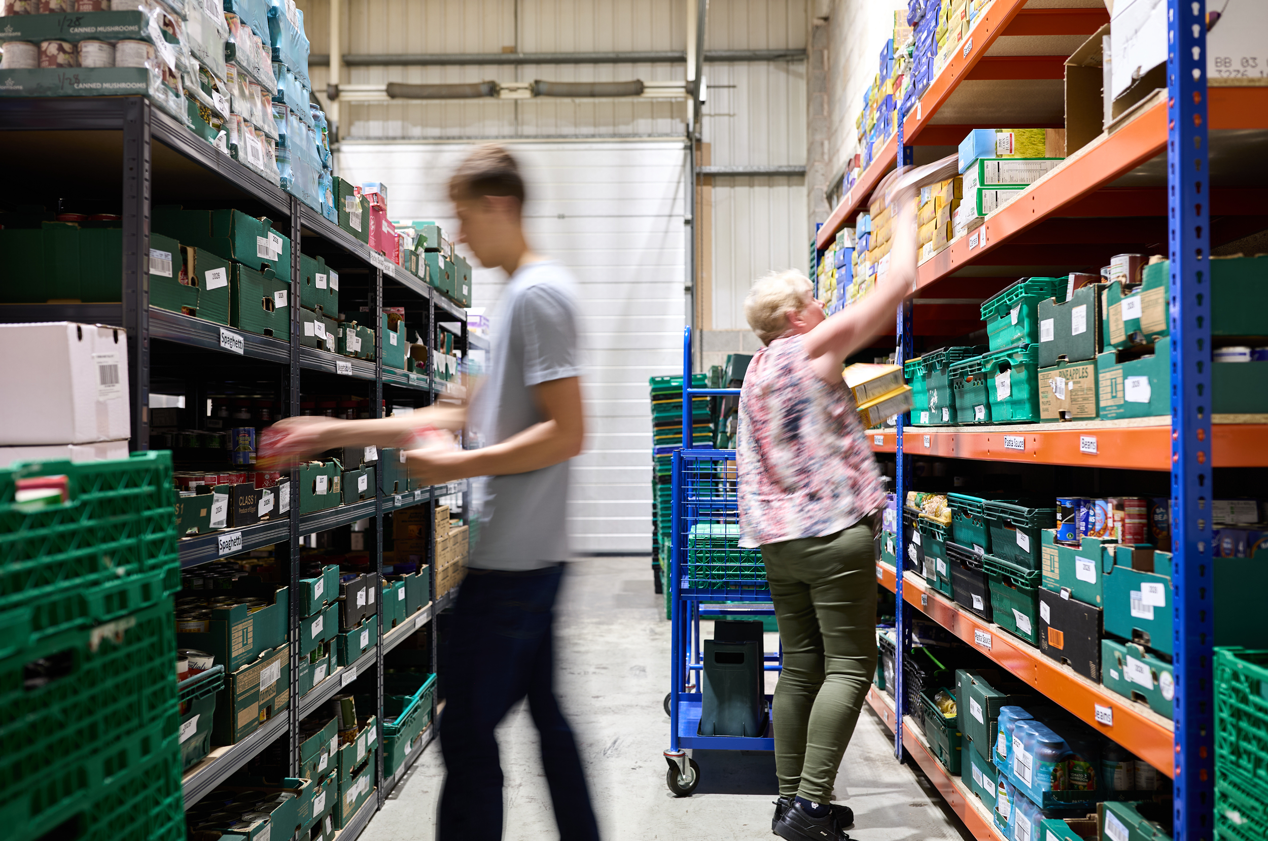 Two people are sorting through the shelves at a foodbank. They are slightly blurry - to show that they are moving quickly around the space.