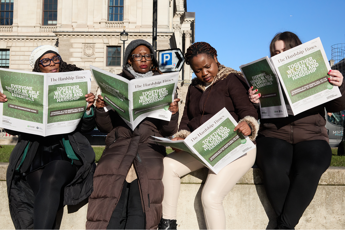 An image of a group of women, sat on a wall. They are all reading the newspaper, which has a green image on the front of it.