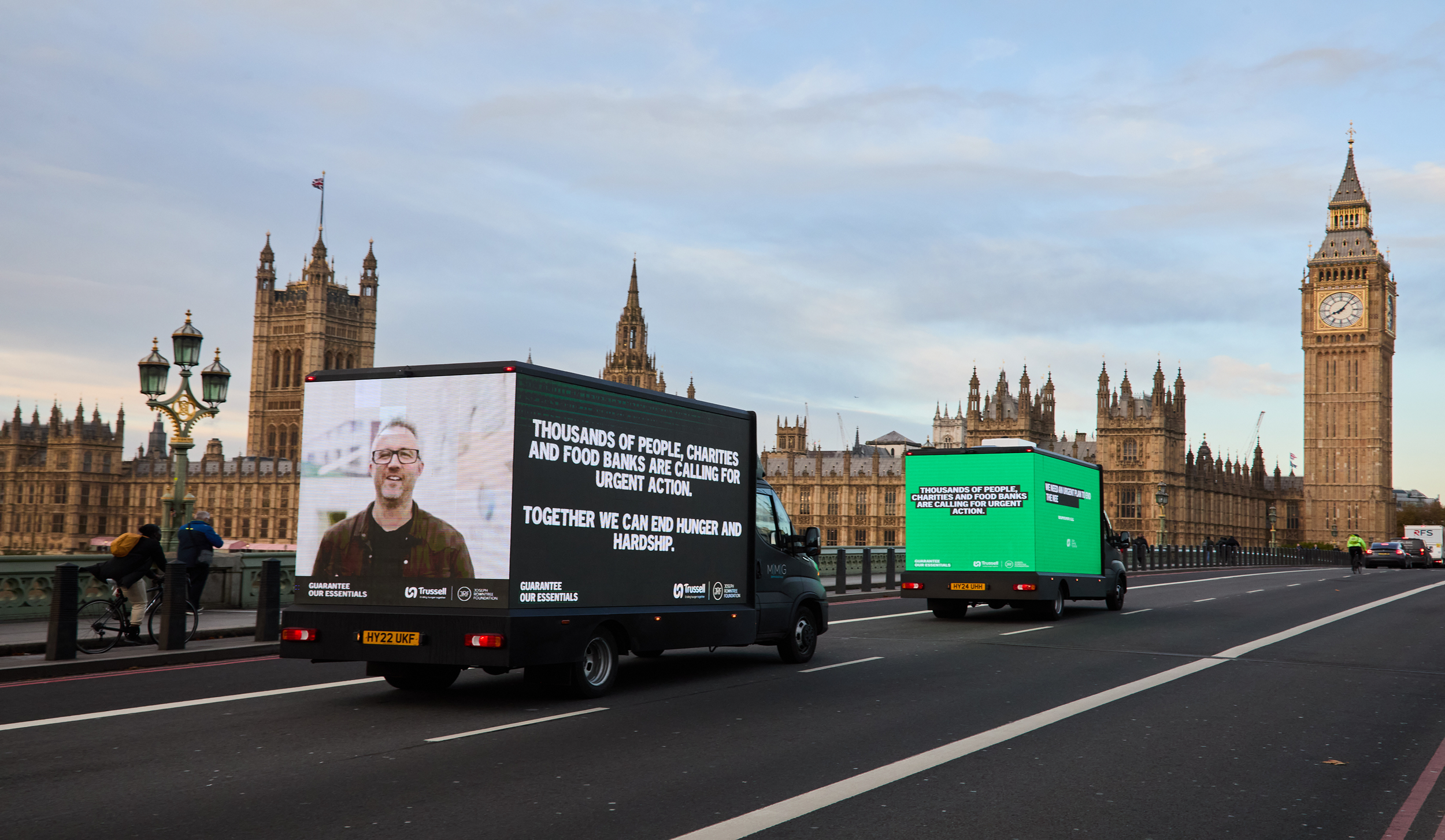Two vans drive over Westminster bridge, towards big ben. They have a video playing on the back of one, and are covered in slogans like 'Together we can end hunger and hardship'