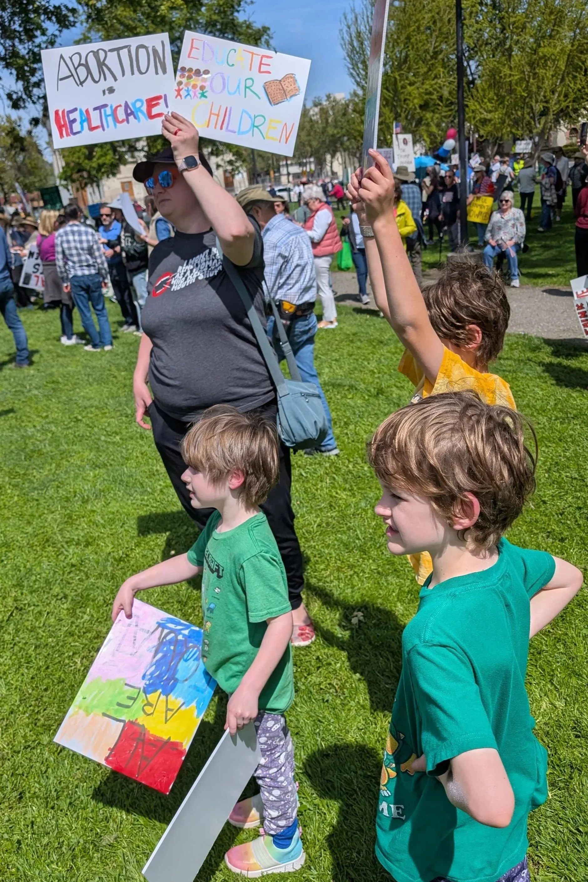 Children and adults participating in a protest outdoors in a park, holding signs with messages related to healthcare and abortion rights, with trees and a crowd in the background.