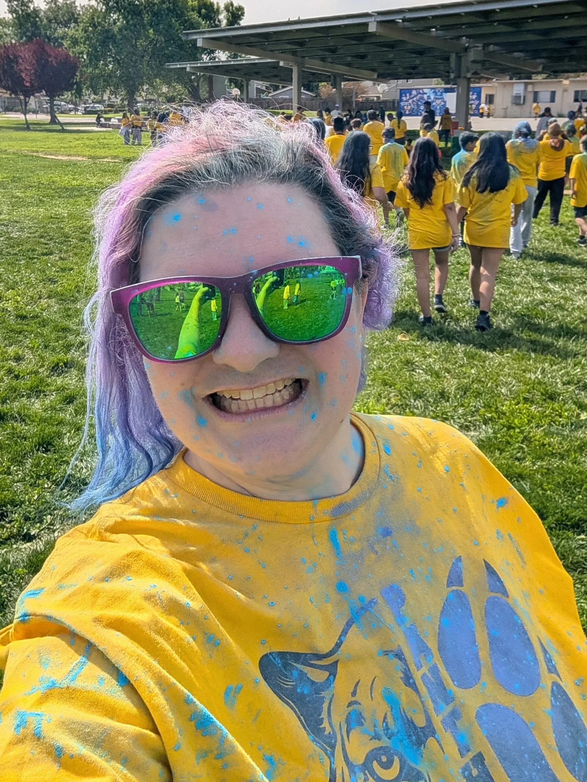 A woman with purple hair and blue chalk splatters taking a selfie at a school during a fun run fundraiser with many people wearing yellow shirts.