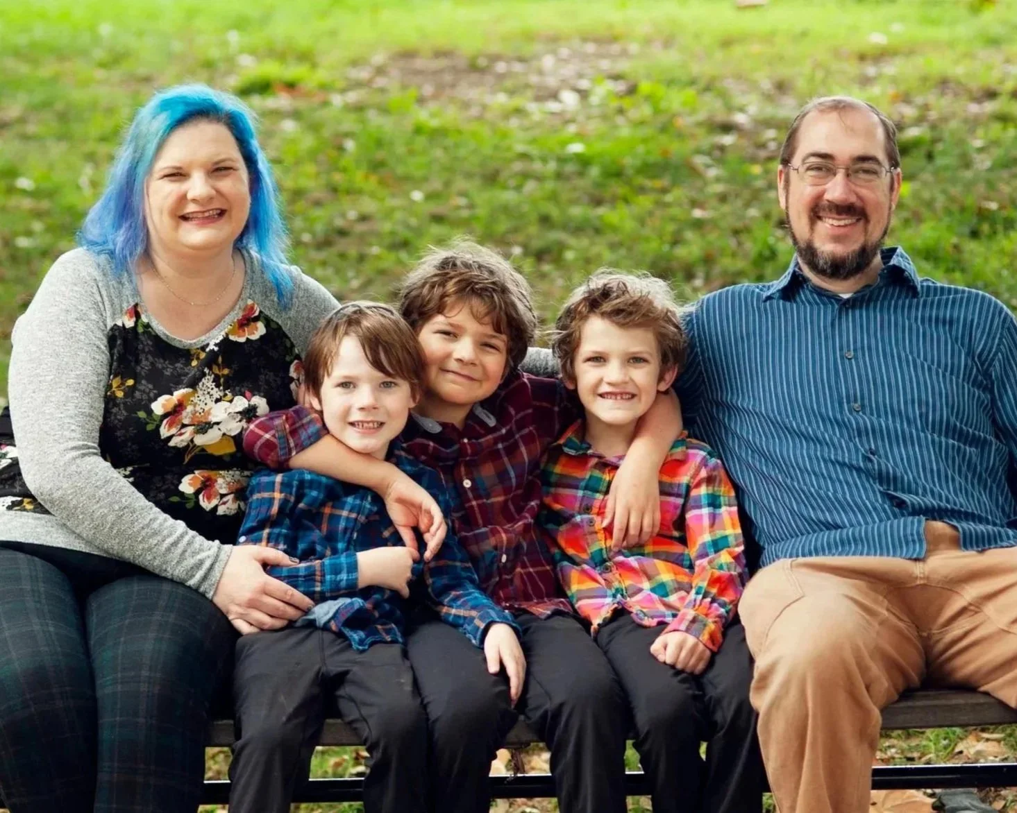 A family of five sitting on a park bench outdoors, smiling at the camera. The group includes two adults and three children, all wearing colorful plaid shirts.