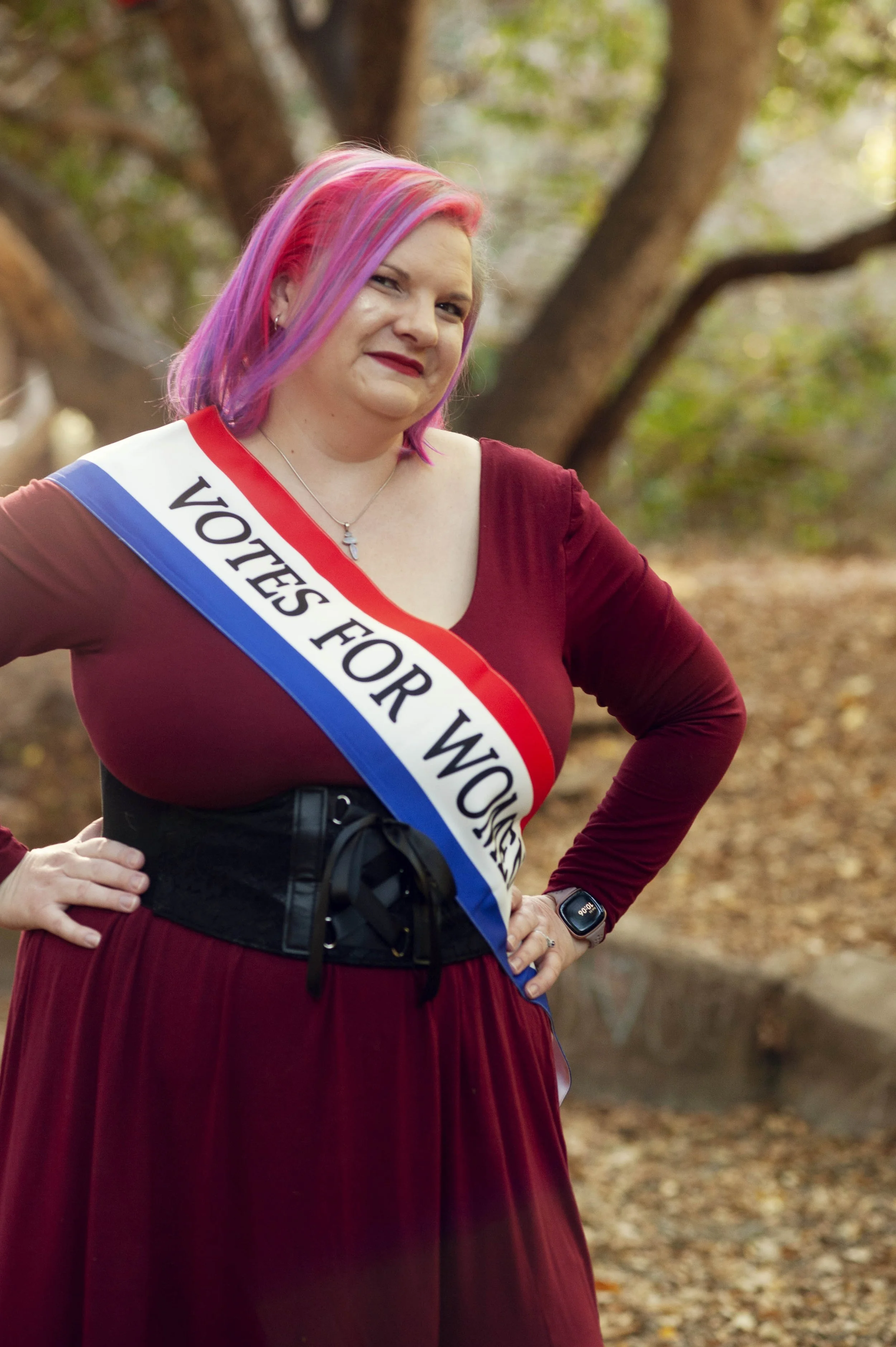A woman with pink and purple hair wearing a burgundy dress and a sash that reads 'VOTES FOR WOMEN' standing outside in a wooded area.