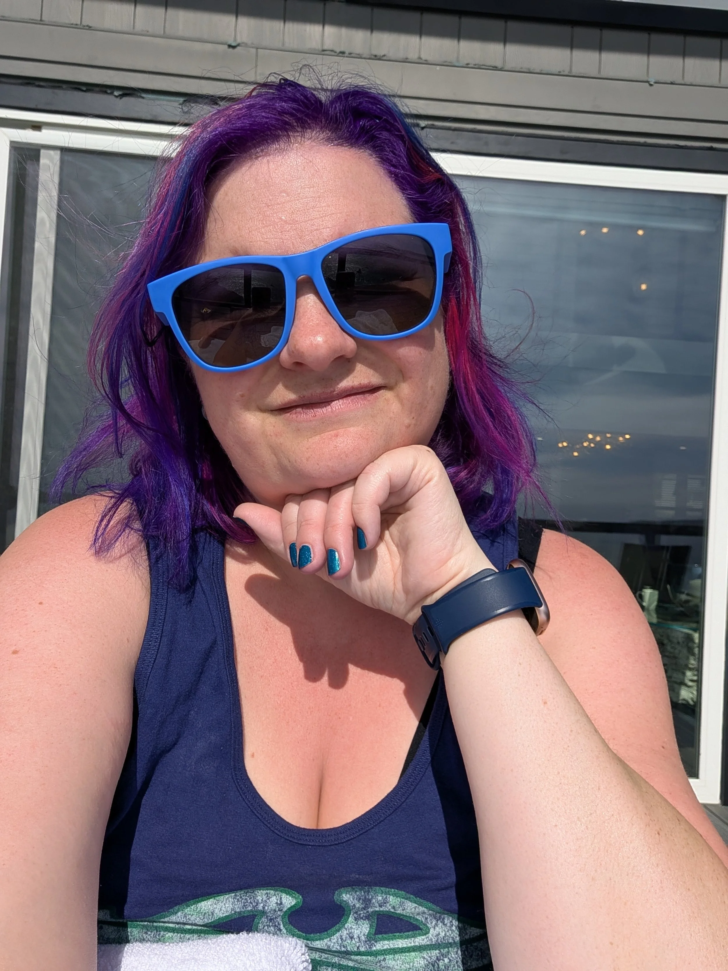 Close-up of a woman with purple and pink hair wearing blue sunglasses, a navy tank top, and a black wristband, sitting outdoors in front of a glass door.