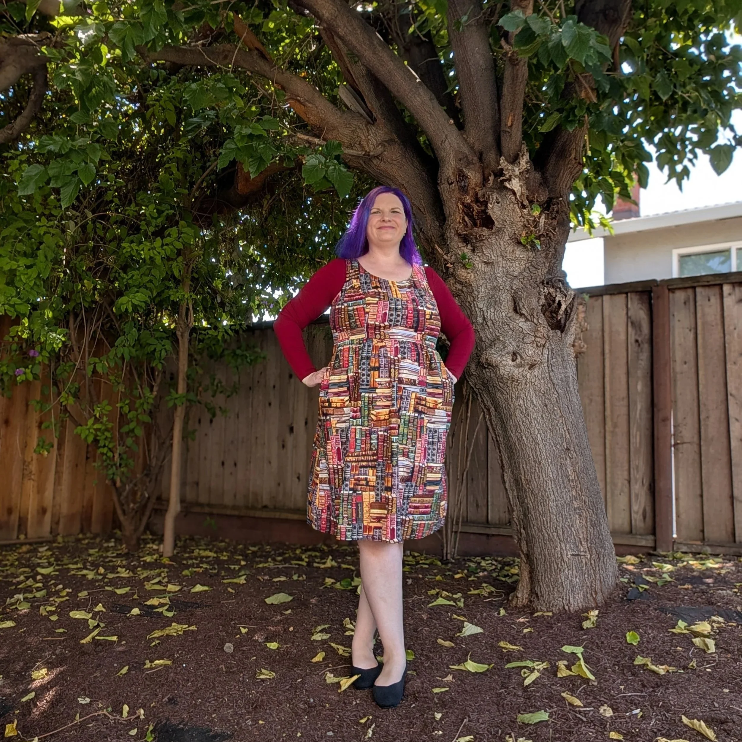 A woman with purple hair smiling and standing with her hands on her hips in front of a large tree with green leaves in a backyard, with fallen leaves on the ground and a wooden fence in the background.