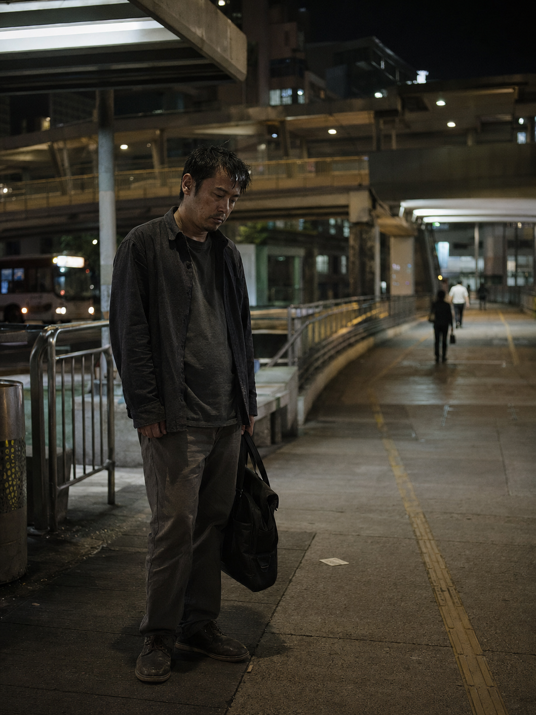 A man standing alone on a city sidewalk at night under an overhead structure, carrying a black duffel bag, with a few other people walking in the background.