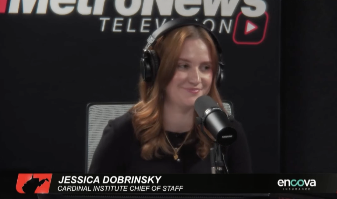 A woman with long red hair wearing headphones and a black shirt, sitting in front of a microphone in a radio studio with a black background. The background has the partial text 'Meter. or News' and a YouTube logo. She is smiling slightly and looking to her left. A name tag indicates she is Jessica Dobrinsky, the Chief of Staff at the Cardinal Institute.