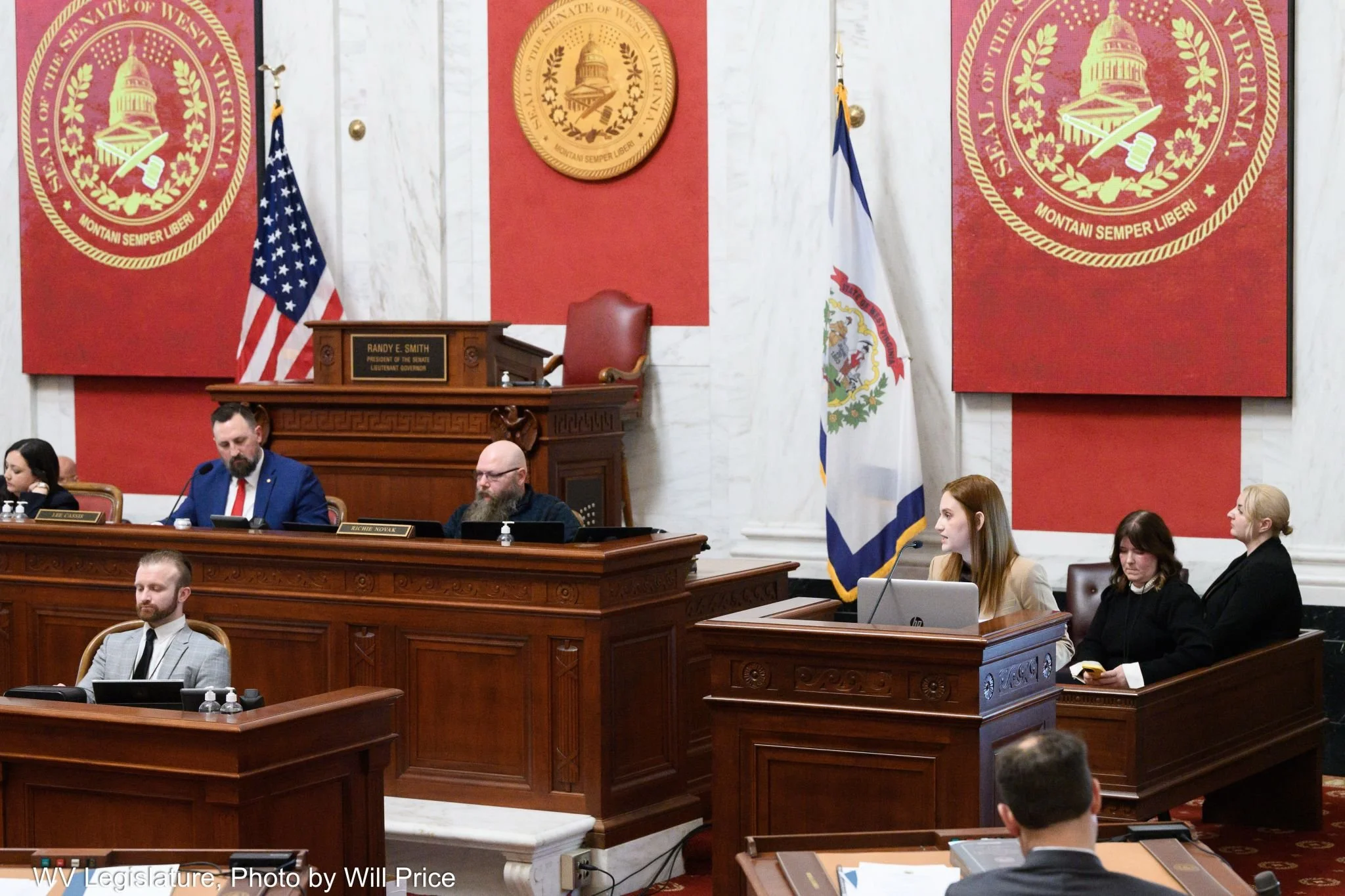 A legislative chamber with people seated at desks, including men and women. Prominent wooden desk at the front has a nameplate for Randy E. Smith, President of the Senate, Lieutenant Governor. Blue and white flags are behind the desk. The background has red adorned panels with gold emblems, one of which features a dome and the phrase "Montani Semper Liberi" indicating West Virginia.