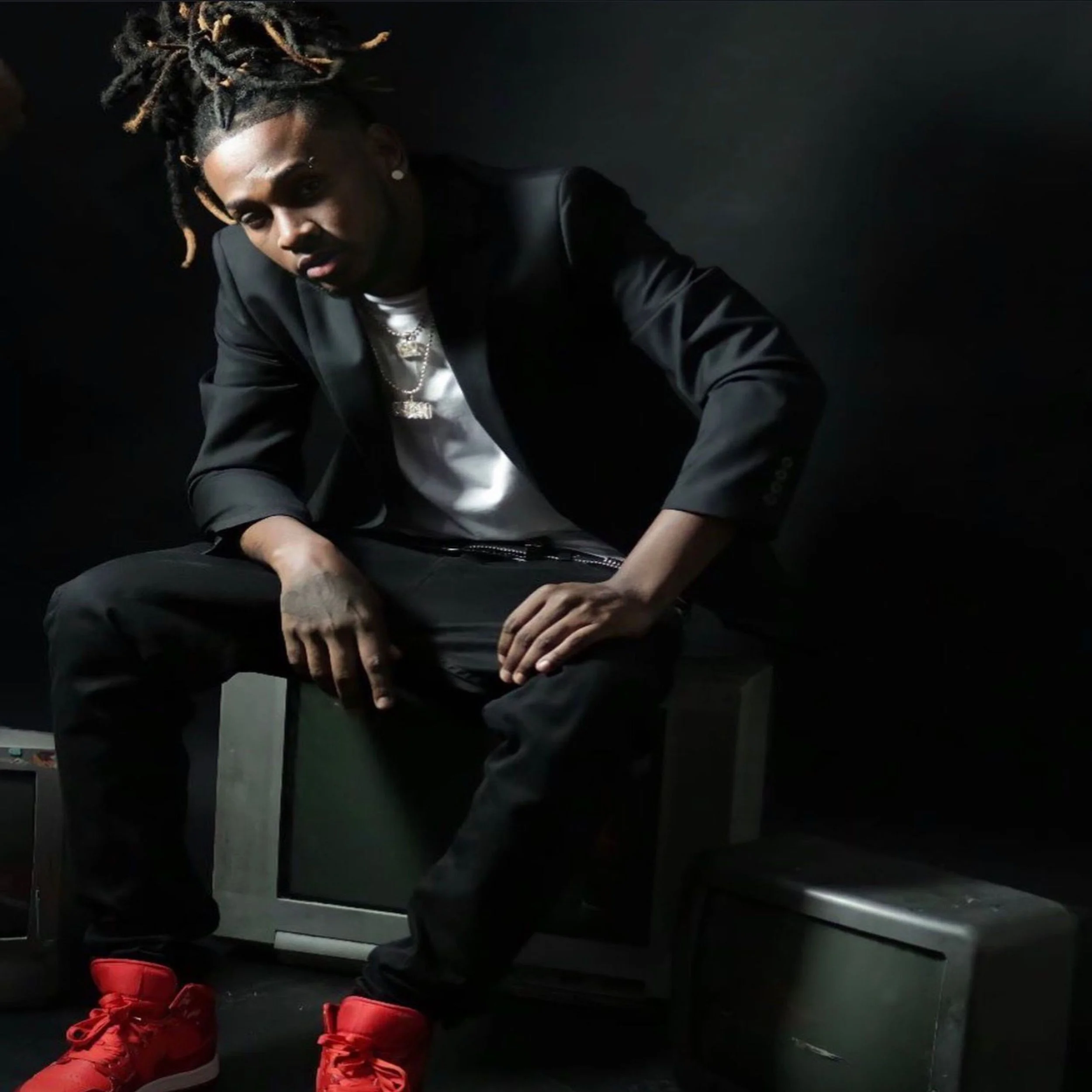 Persona con peinado de dreadlocks rubios, ropa oscura, relojes y cadenas, en un fondo oscuro, con zapatos deportivos rojos en primer plano.