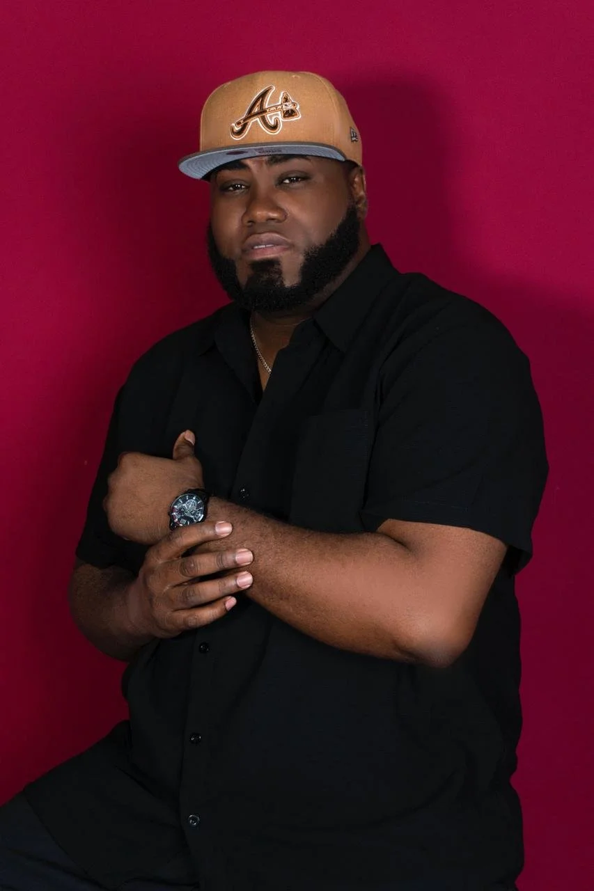 Hombre con gorra de cola de tigre color oro, camiseta negra, reloj en la muñeca izquierda y fondo rosa.