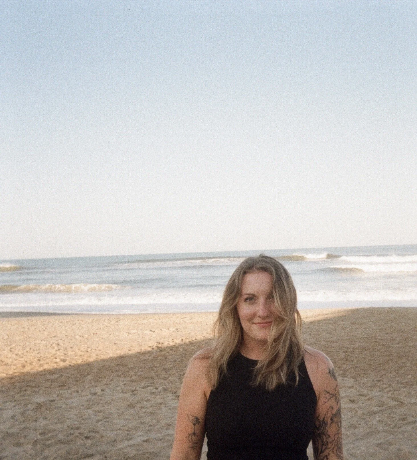 A woman with long wavy blonde hair standing on a sandy beach near the ocean, smiling at the camera, with waves in the background under a clear sky.