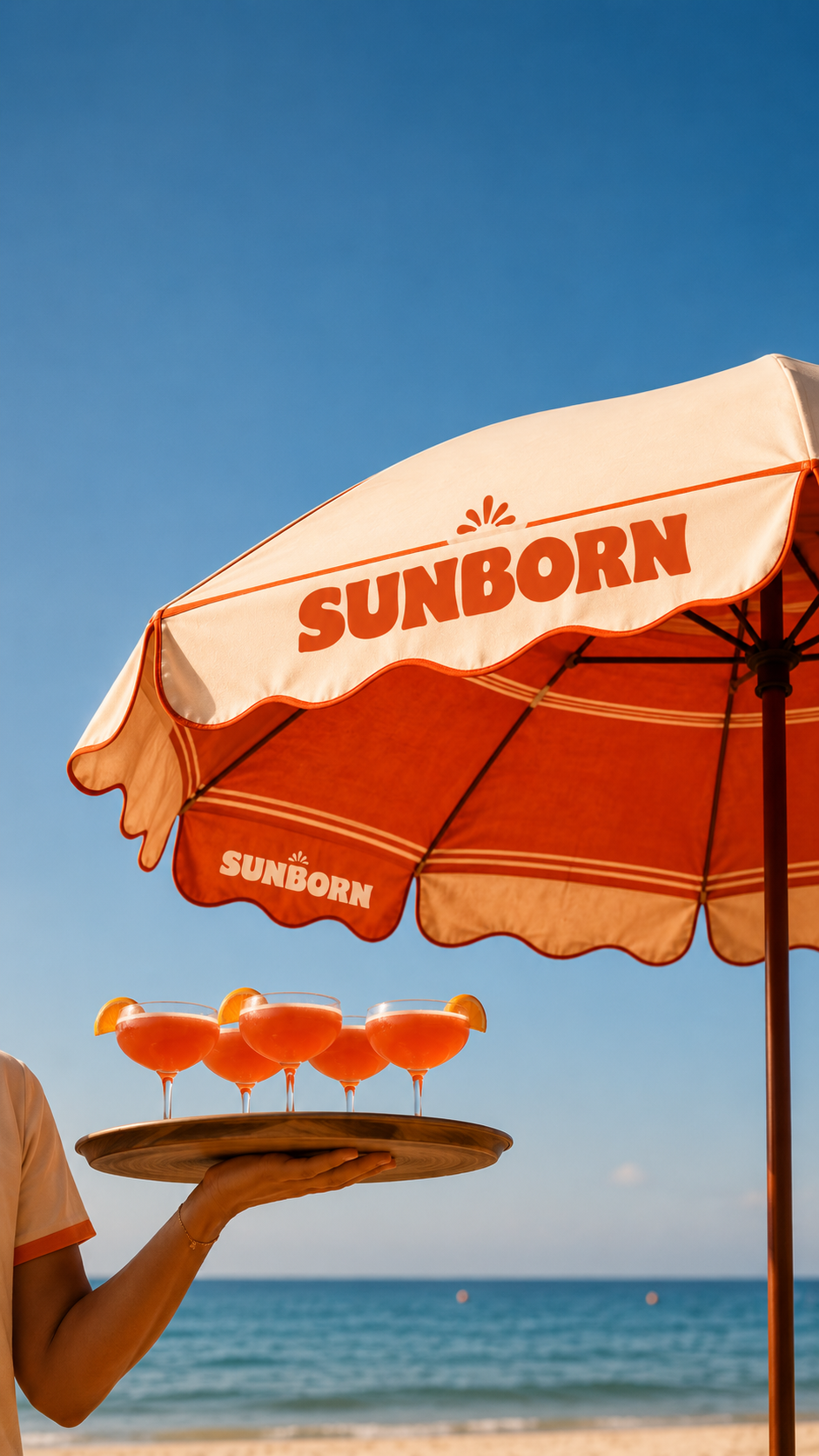 Four pink cocktails with orange garnishes on a wooden tray, held by a woman at the beach under an orange Sunborn umbrella.