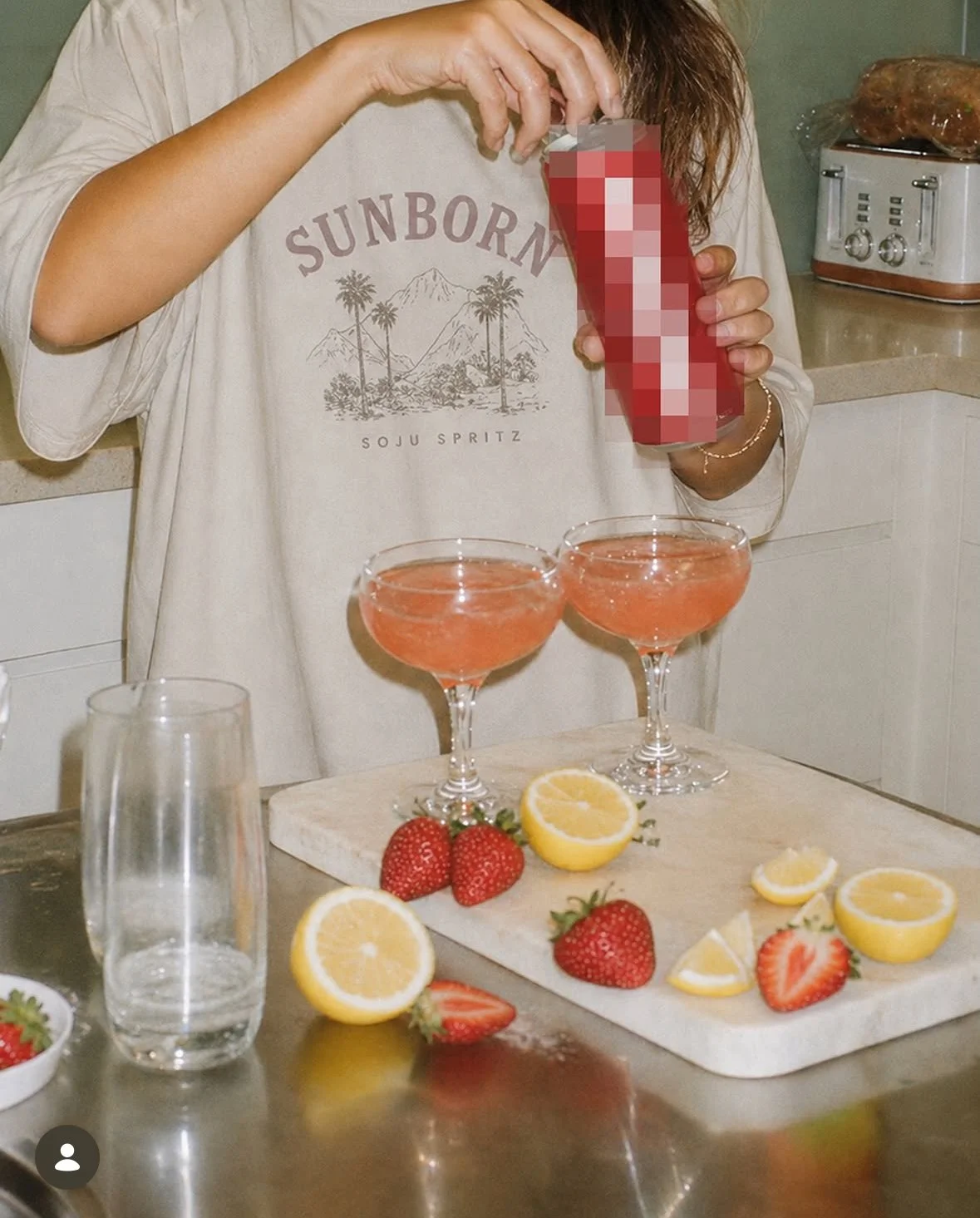 A person is preparing two pink cocktails with strawberries and lemon slices on a kitchen counter. There are fresh strawberries and lemon halves, slices, and lemon wedges on a cutting board, along with a clear glass of water and a small bowl of strawberries. In the background, a toaster oven and a plastic-wrapped loaf of bread are visible.