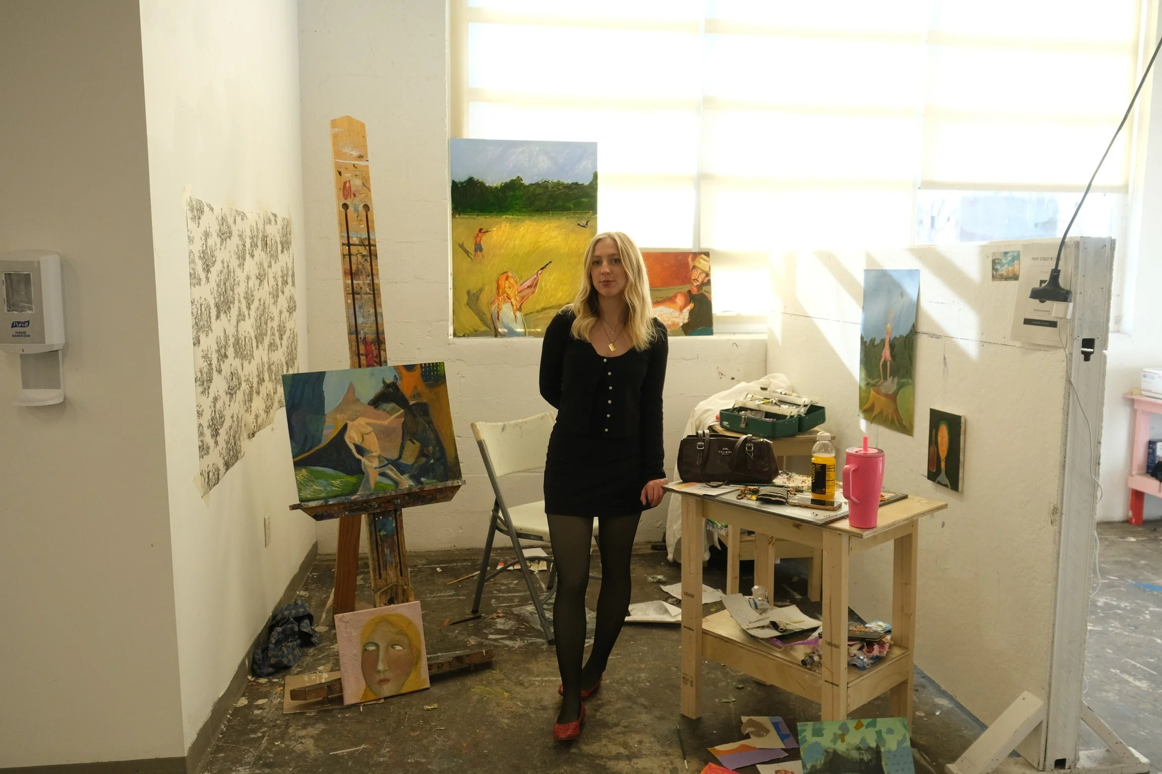 A woman standing in an art studio surrounded by paintings and art supplies, with sunlight streaming through a large window.