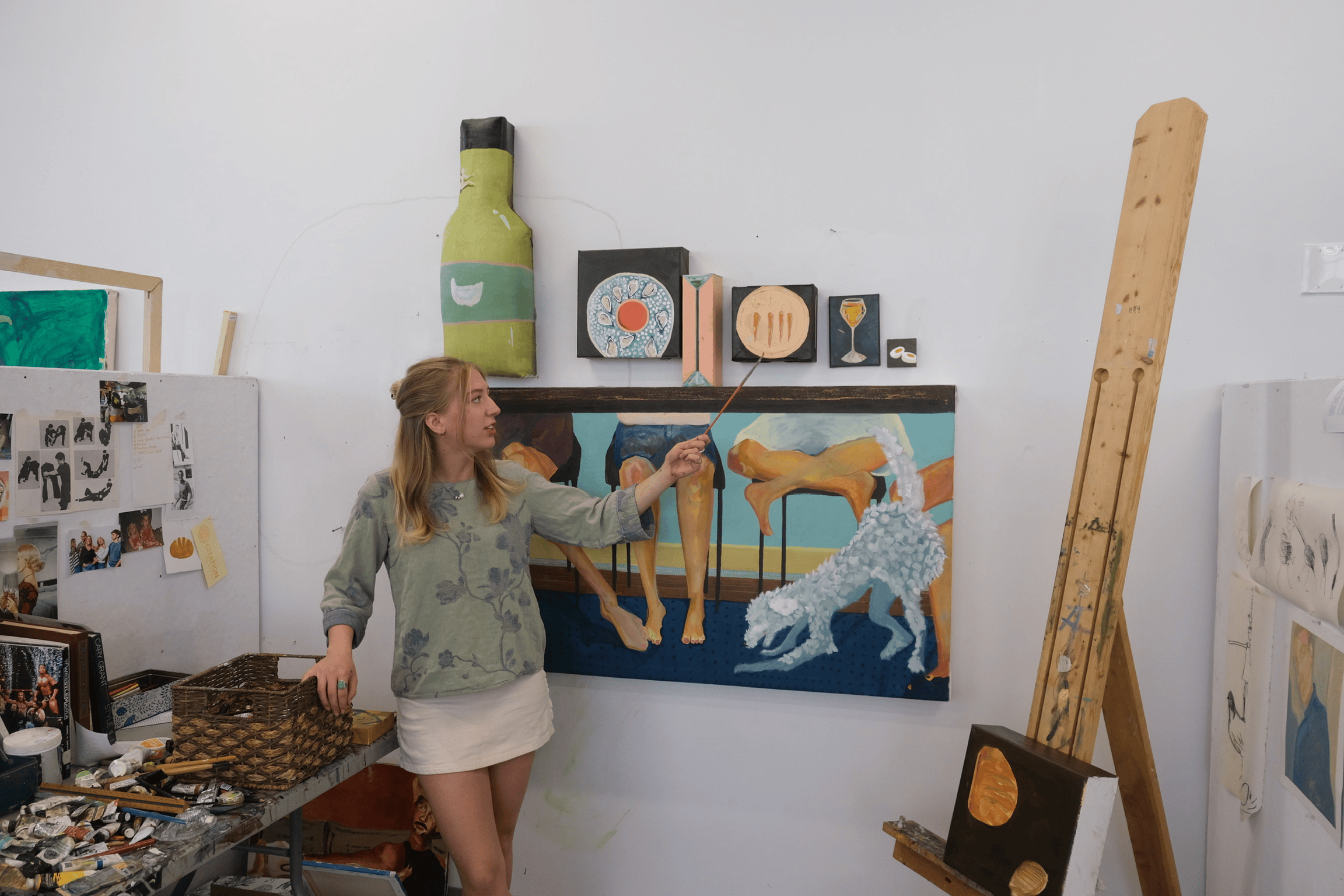A woman in a gray patterned hoodie and white skirt points at a blue painting of a lion on the wall of an art studio. The studio walls display various artworks, including painted plates and food-themed pieces. There is an easel with a partially painted canvas on the right.