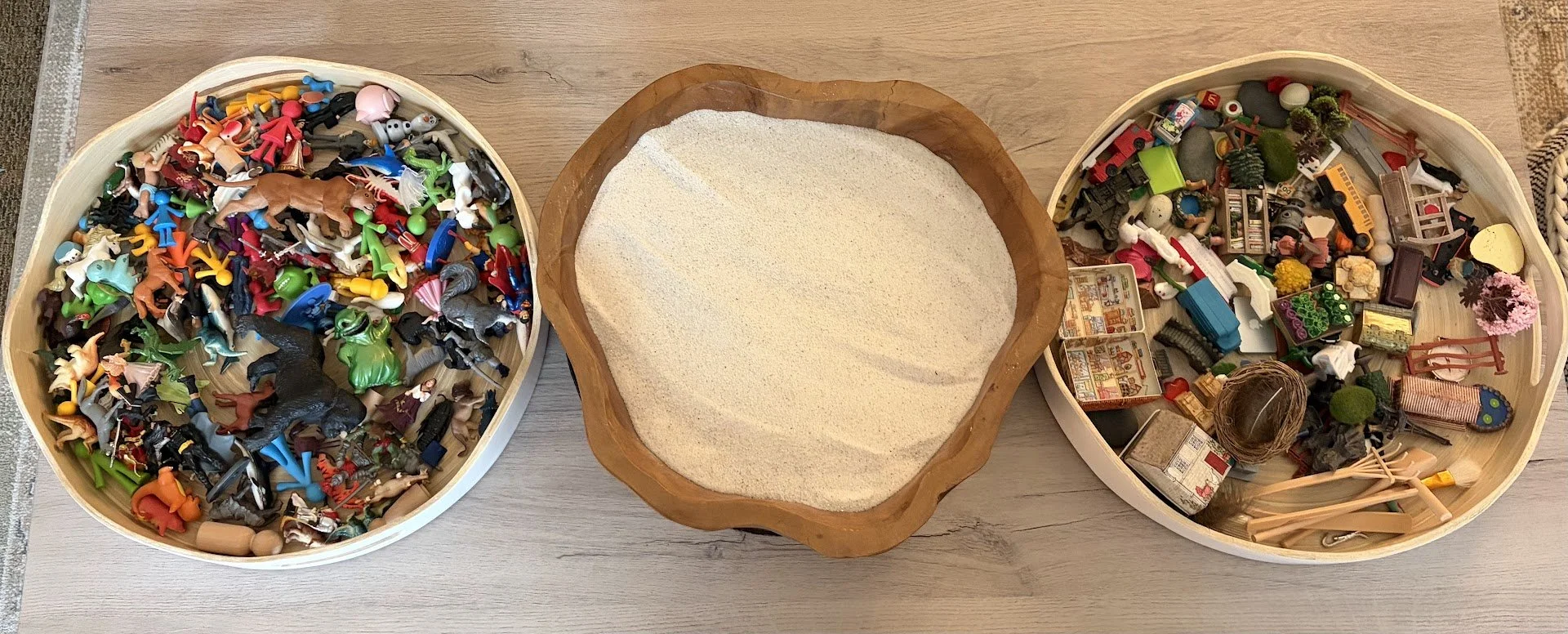 Three baskets filled with small toys, sand tray in the middle, and toys and decorations around, on a wooden surface.