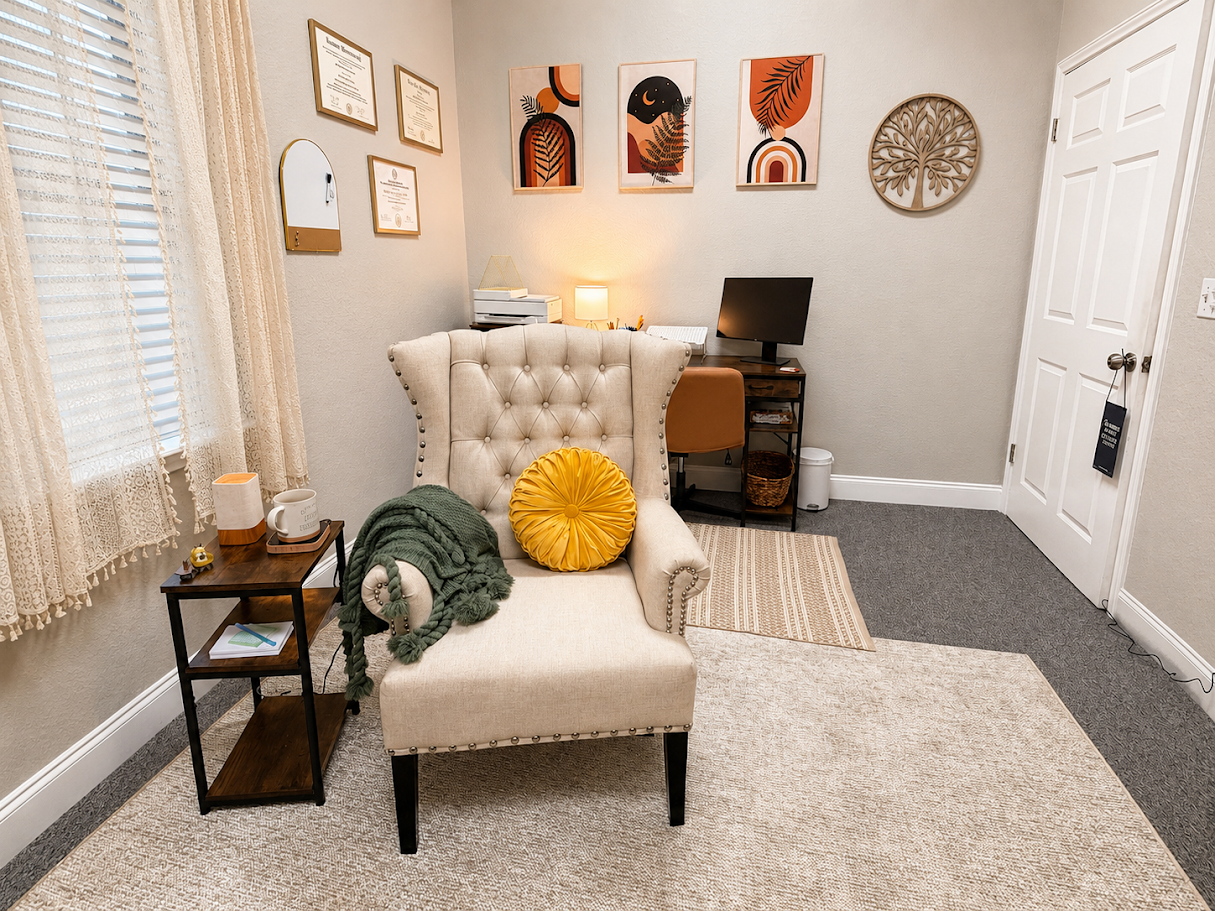 A cozy home office with a beige tufted armchair in the center, a green throw blanket and a yellow round pillow on it, and a small side table with a mug, tissue box, and decor. Behind the chair, there are framed documents and colorful artwork, a desk with a computer, and a white door.