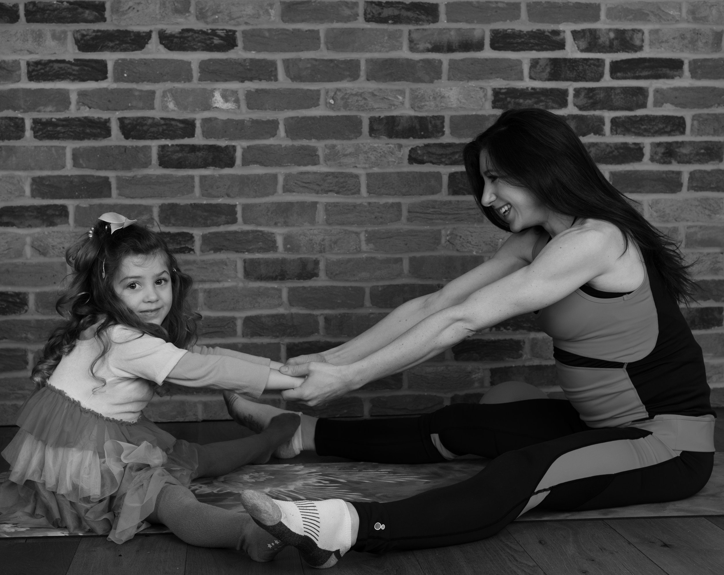 A woman and a young girl are engaging in a playful stretching exercise against a brick wall, holding hands and smiling.