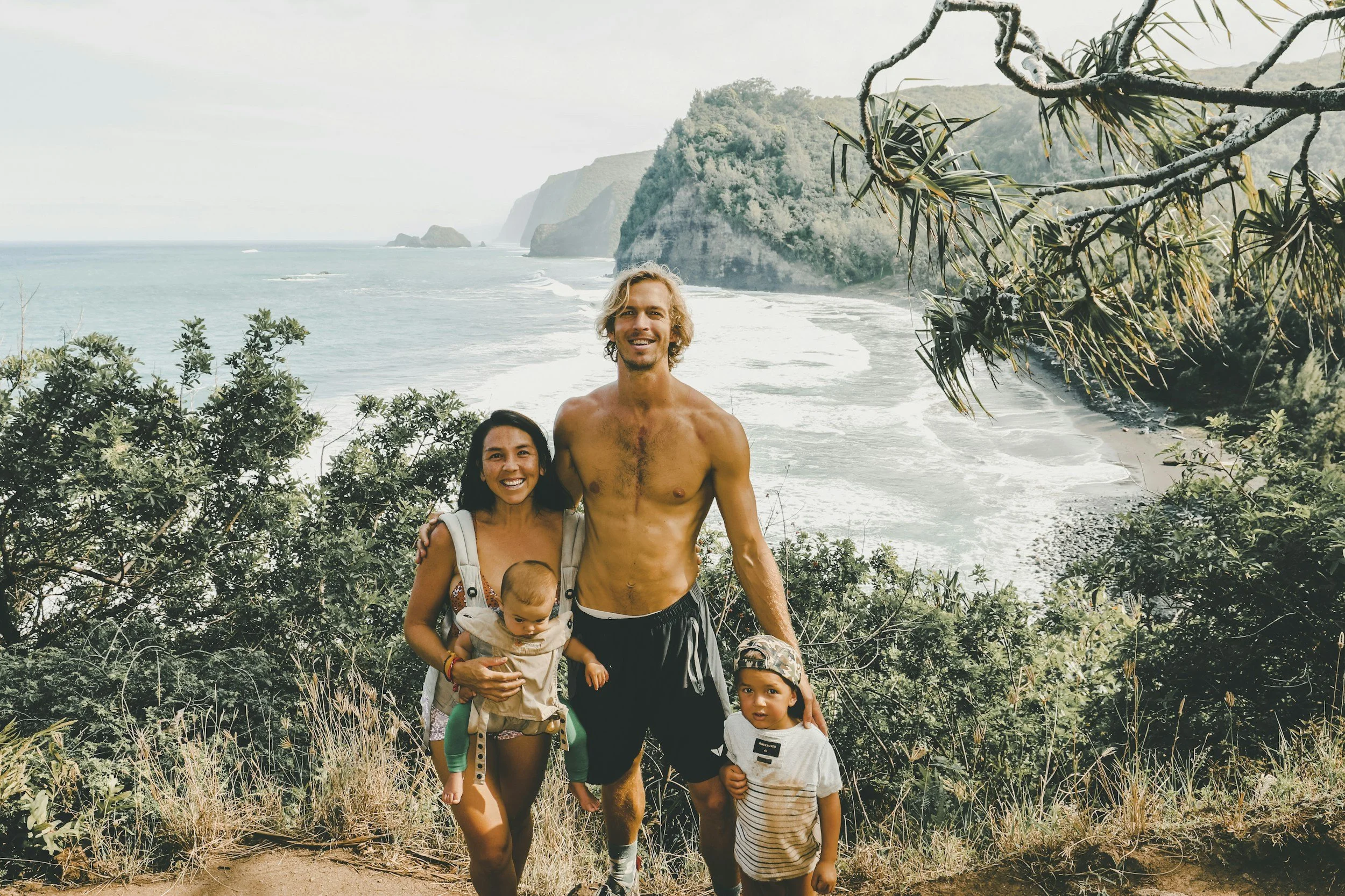 A group of five people, including adults and children, standing on a trail near a coastal area with cliffs, trees, and the ocean in the background.