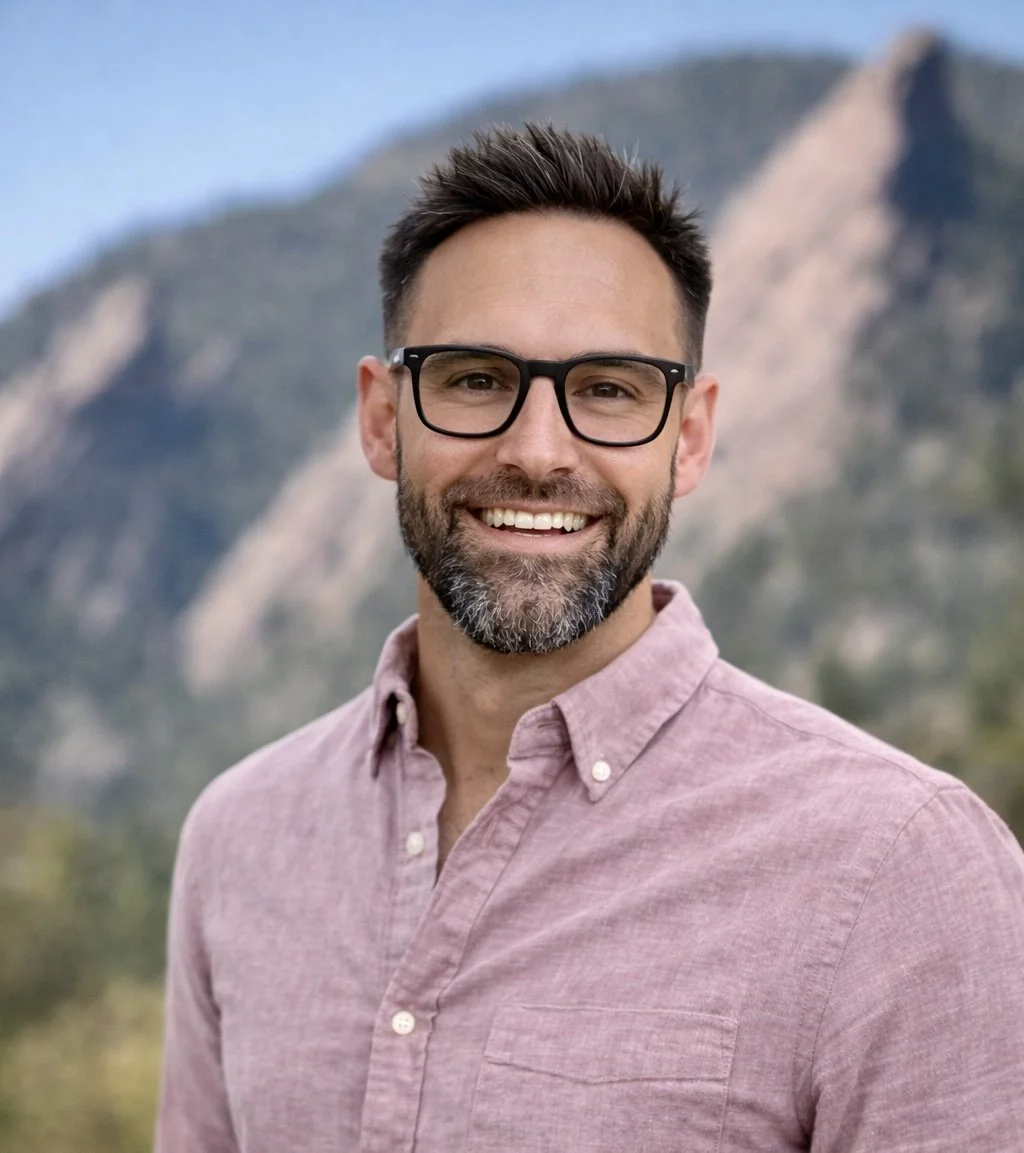 A smiling man with glasses and a beard wearing a pink button-up shirt outdoors with mountainous terrain in the background.