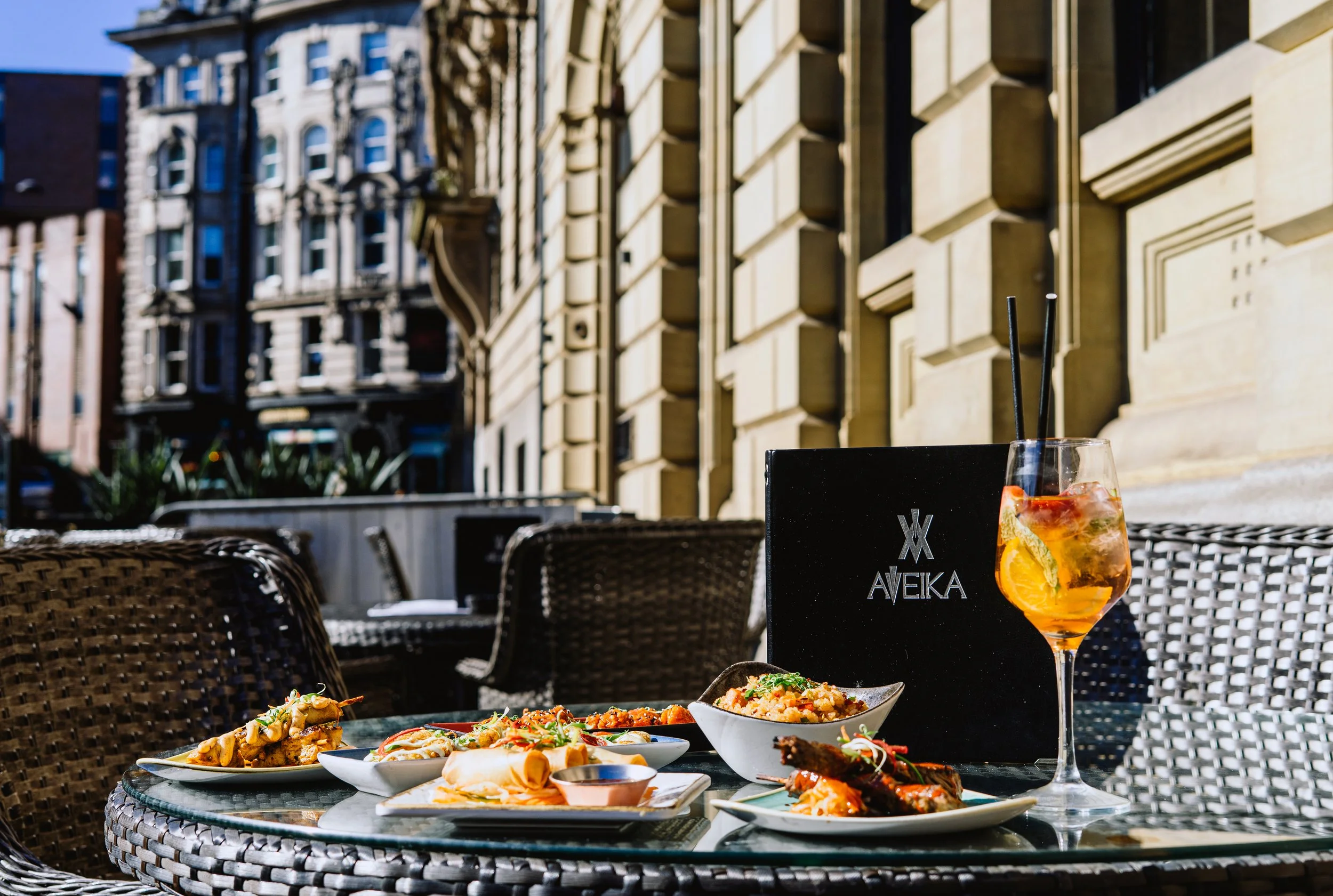 Outdoor restaurant table with various dishes including pasta, rice, and grilled meats, a drink with lemon and mint, and chopsticks, with buildings in the background.