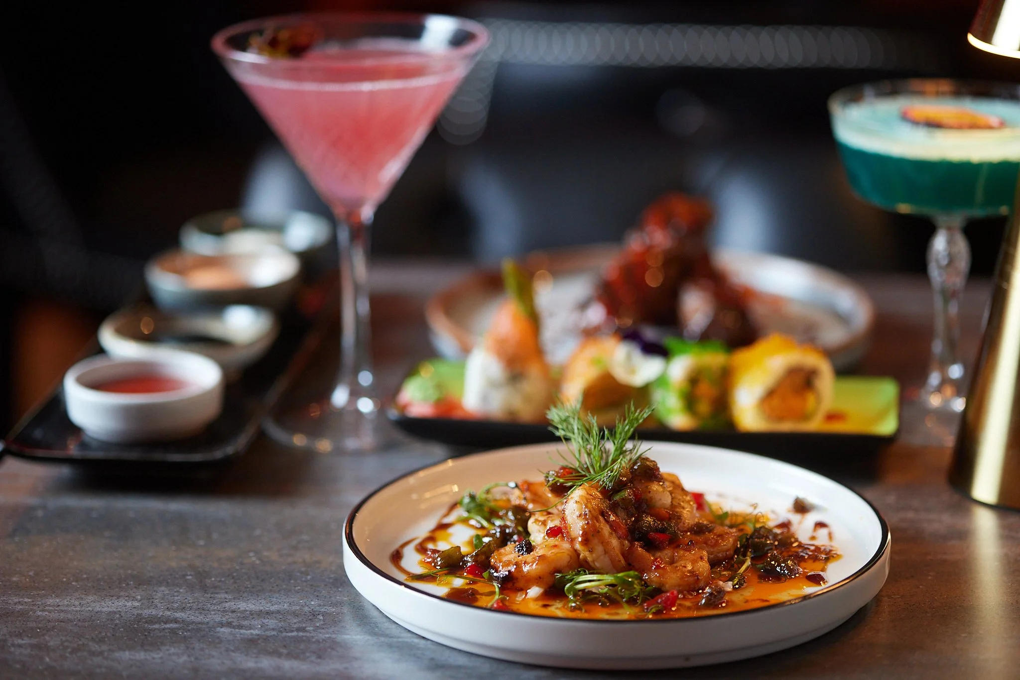 A close-up of a plated dish with seafood and sauce, in the foreground, with various colorful cocktails and small bowls in the background on a dark table.