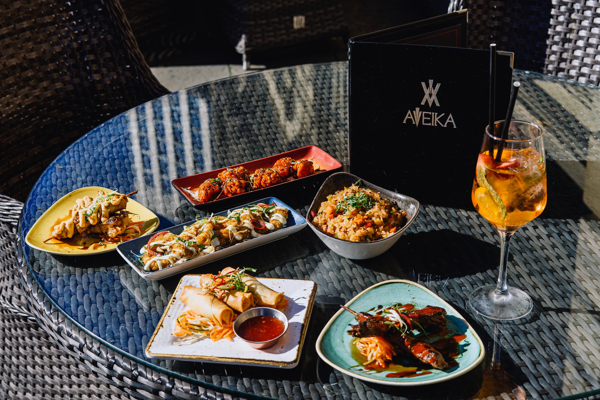 Assorted Asian dishes on a glass table with a drink in a glass and a black menu.
