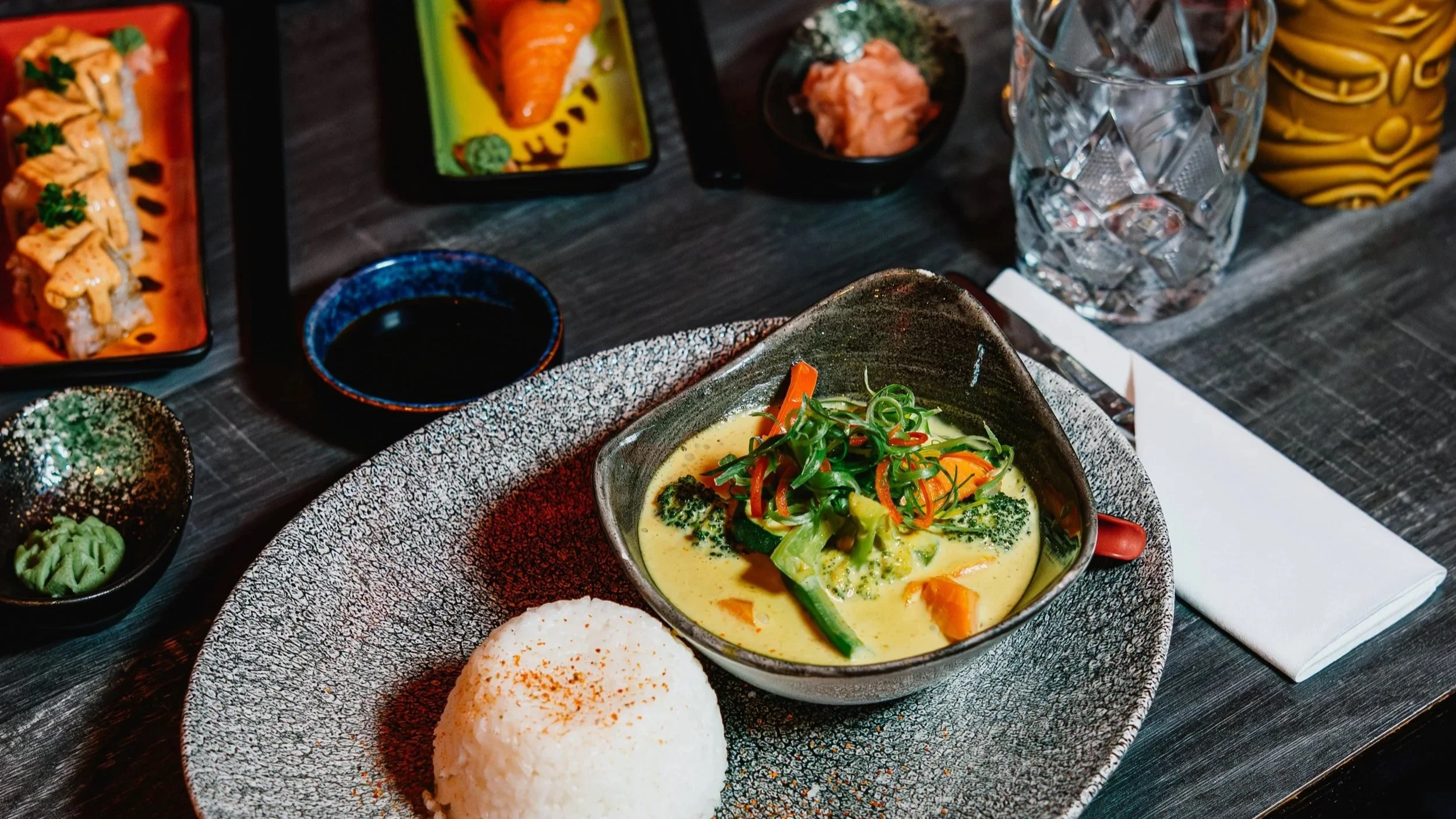A table with various Asian dishes, including a bowl of yellow curry with vegetables and green onions, a mound of steamed white rice, and assorted sushi and side dishes, set on a dark dining table with glasses of ice water.