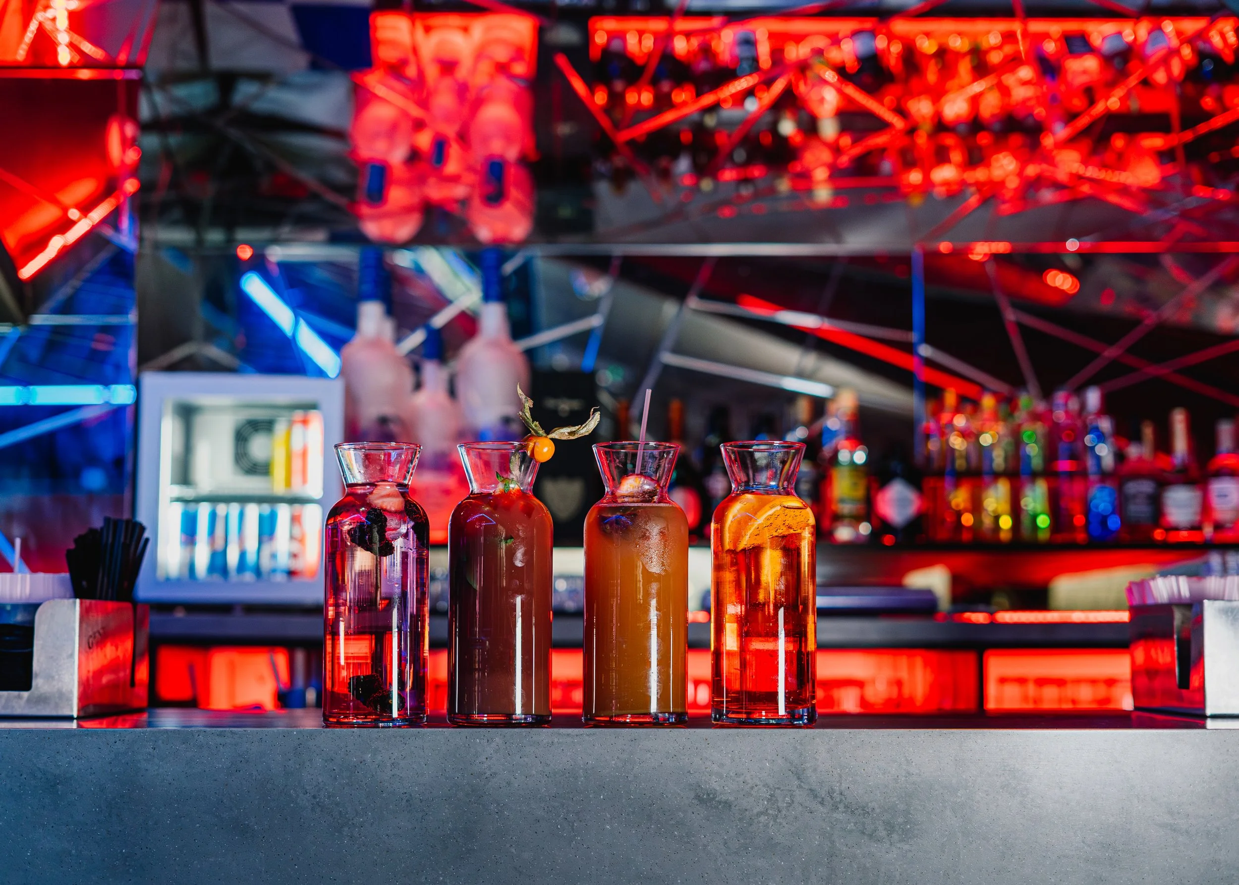 A row of four colorful cocktails with garnishes on a bar counter, with a neon-lit bar in the background.