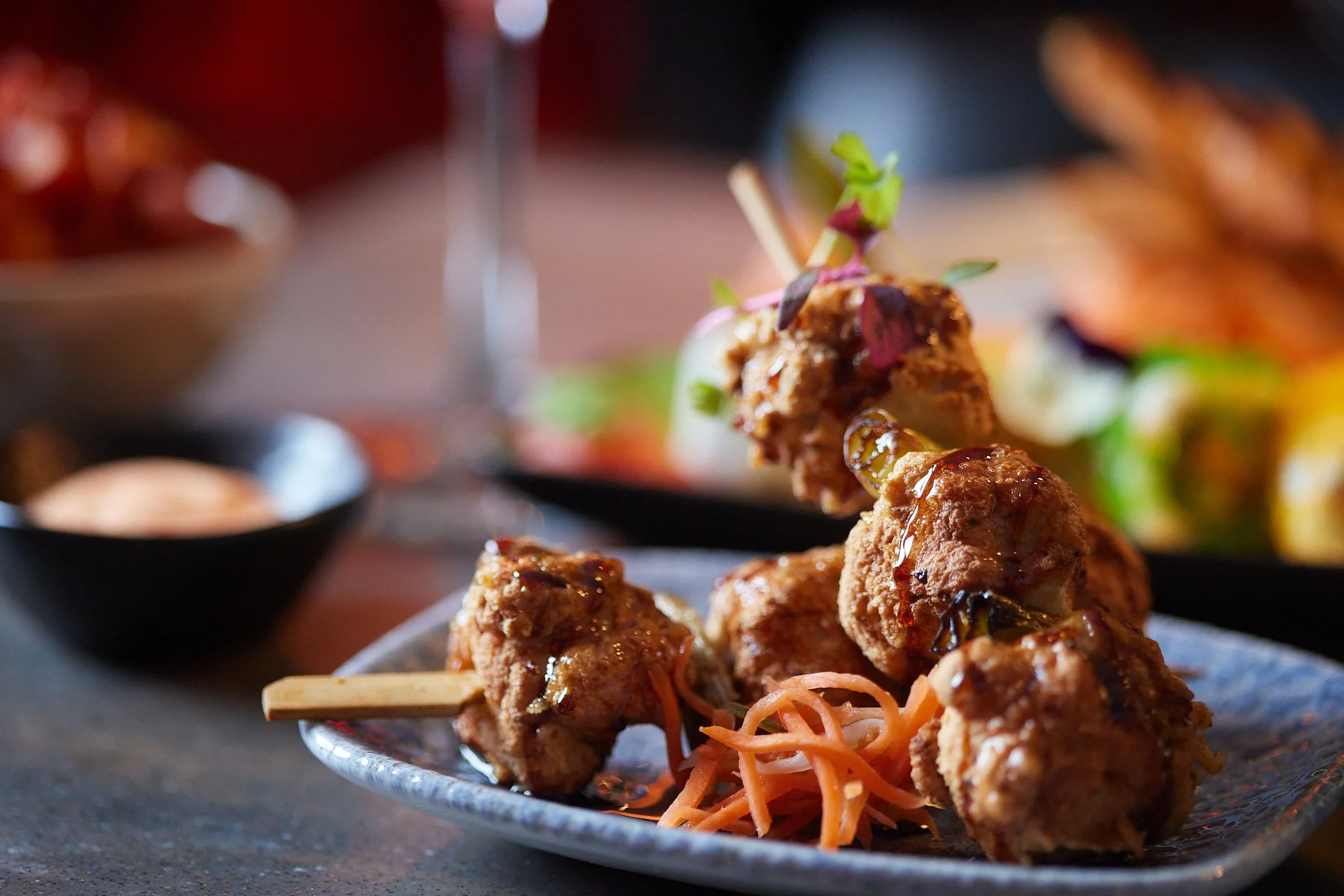 Close-up of skewered meatballs glazed with sauce, garnished with small green herbs, served on a plate with shredded carrots.