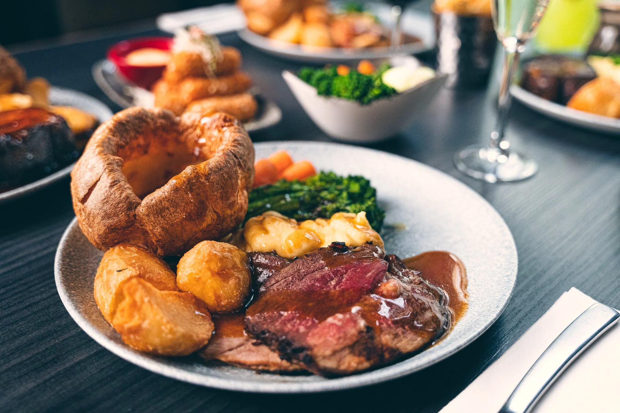 A plate of plated roast beef with gravy, roasted potatoes, and vegetables, including carrots and broccoli, with other dishes and drinks visible in the background.