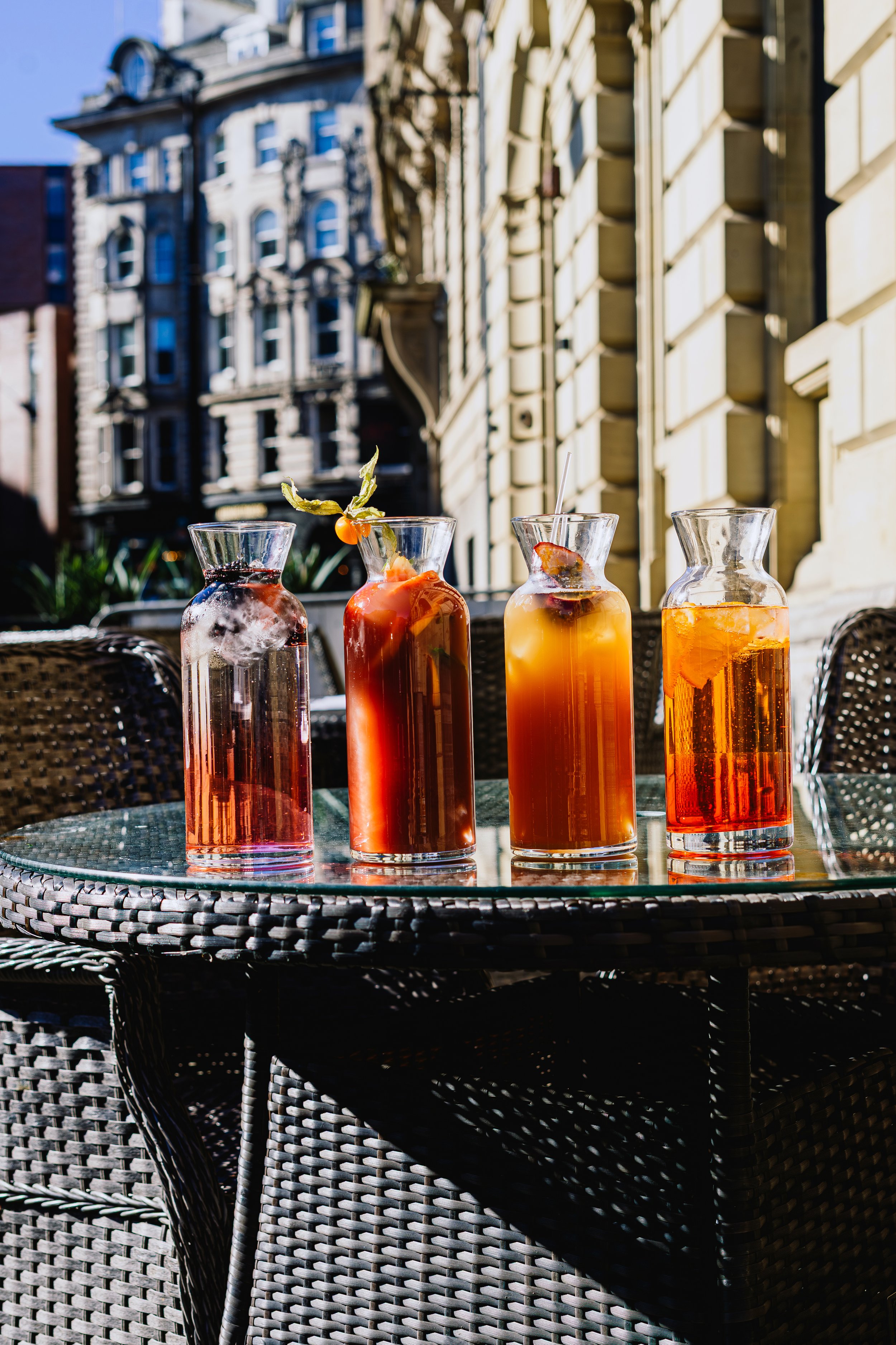 Four glass jars containing colorful beverages arranged on a glass table outdoors, with a wicker chair and buildings in the background.