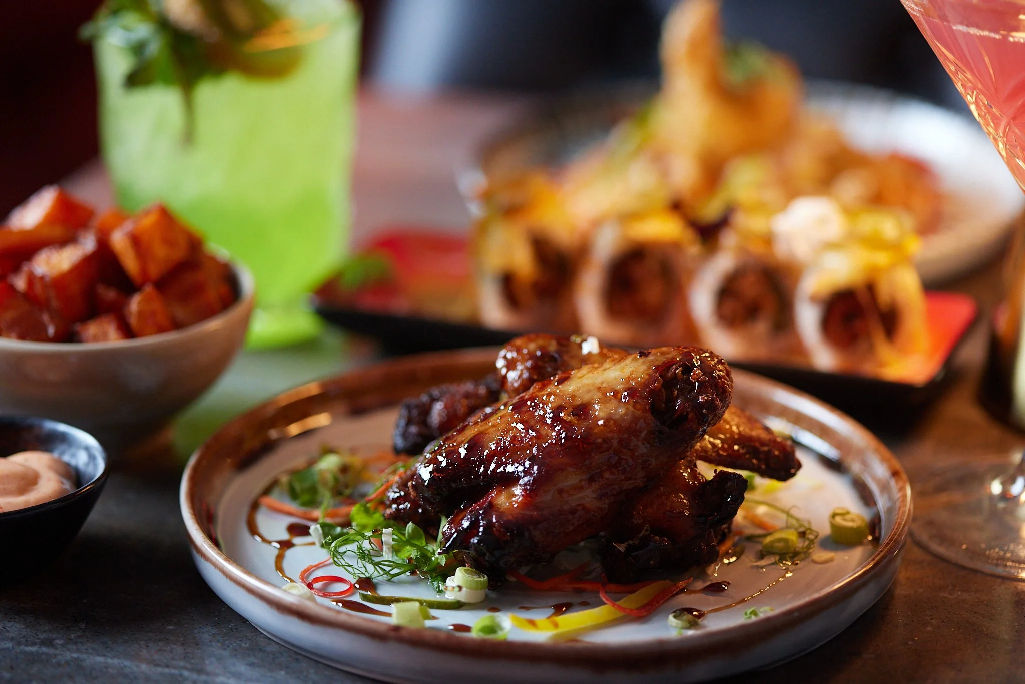 Close-up of a plate with barbecued chicken wing garnished with sliced green onions and herbs, with other dishes and a large tortilla in the background.