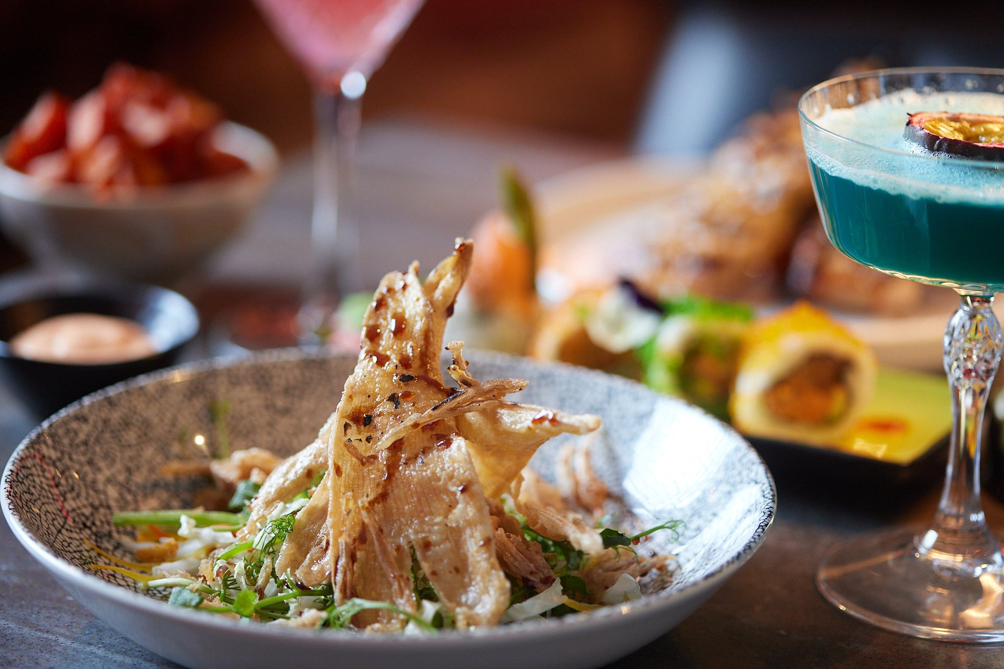 Close-up of Asian-style fried chicken served on a bed of shredded vegetables in a decorative bowl, with a layered blue cocktail garnished with a citrus slice and berry in a cocktail glass in the background.