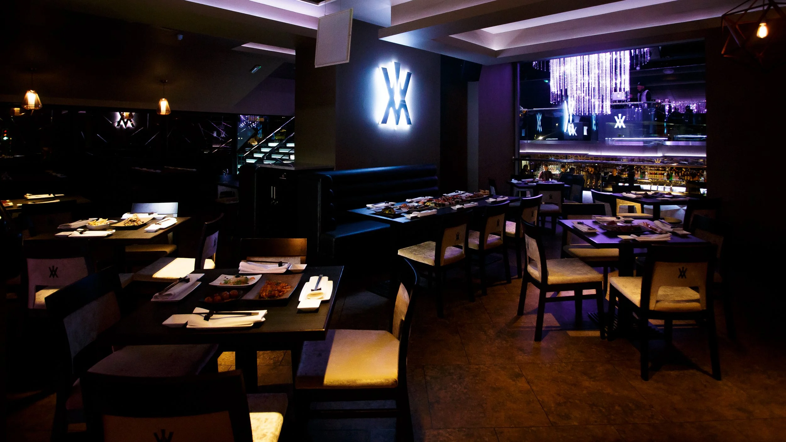 A dimly lit restaurant with tables set for dining, some with plates of food. There are decorative light fixtures, a bar area with bottles visible, and illuminated signage with a stylized 'W' on the wall. The atmosphere appears modern and upscale.