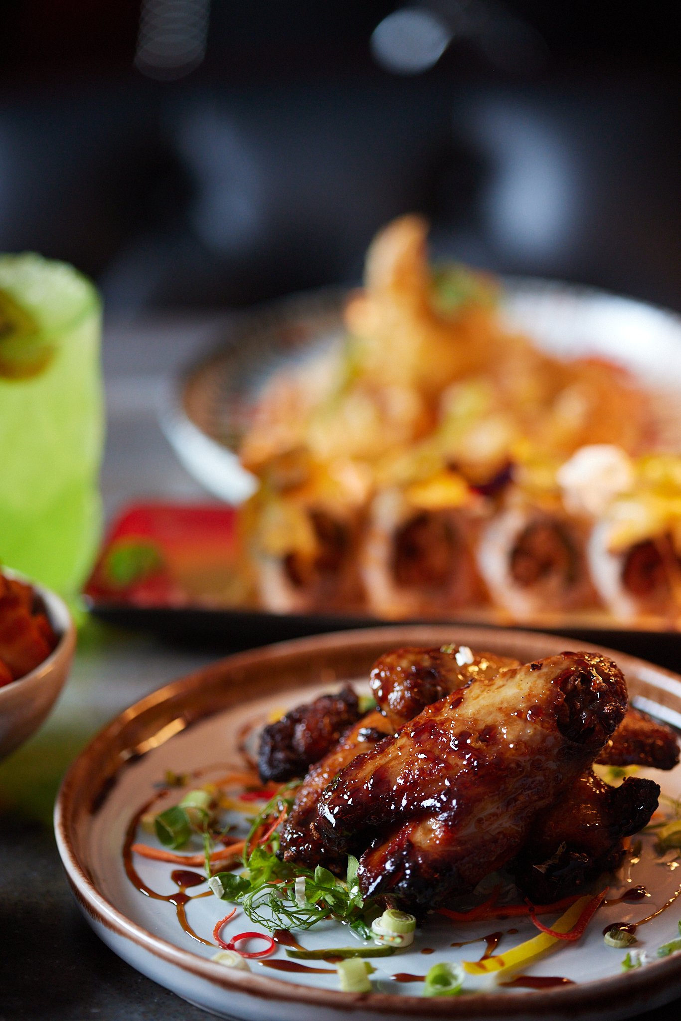 Close-up of a glazed roasted chicken leg on a decorative plate garnished with green onions and microgreens, with a background of various dishes and dim lighting.