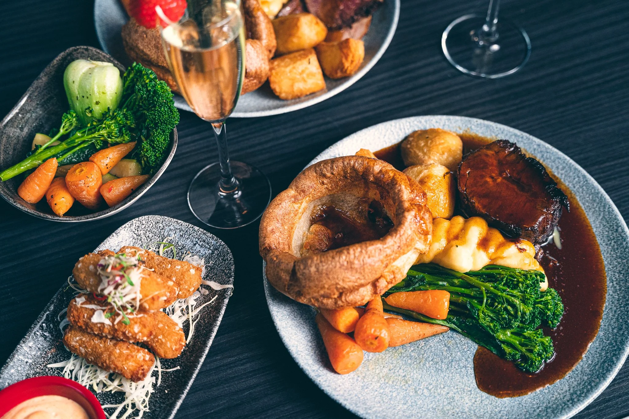 A variety of dishes on a dark table, including roasted vegetables, meat with gravy, a bread bowl with food, and a fried item, along with a glass of champagne with a strawberry garnish and a glass of red wine.