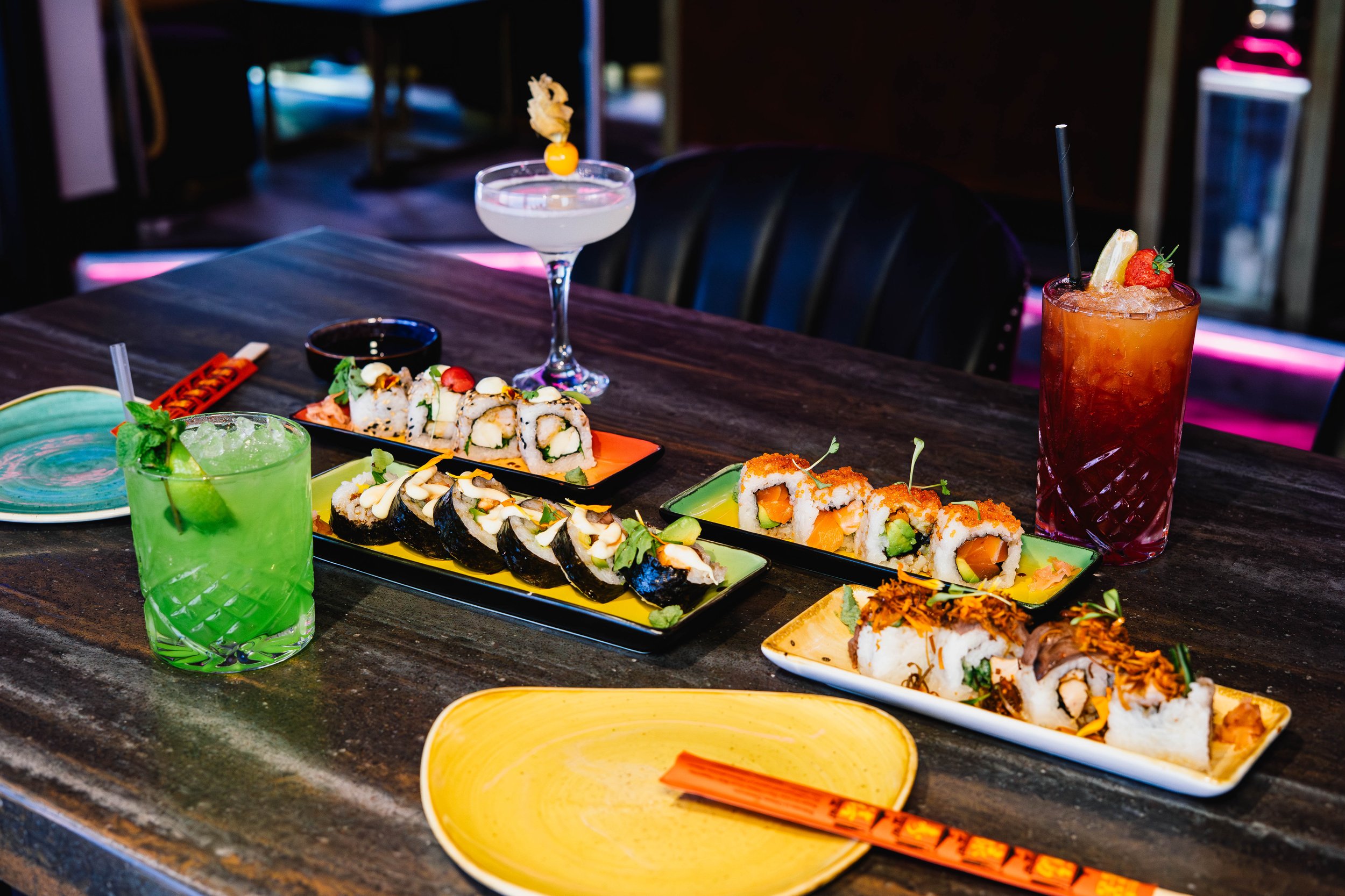 A variety of sushi rolls, cocktails, and small plates on a dark wooden table in a restaurant setting.