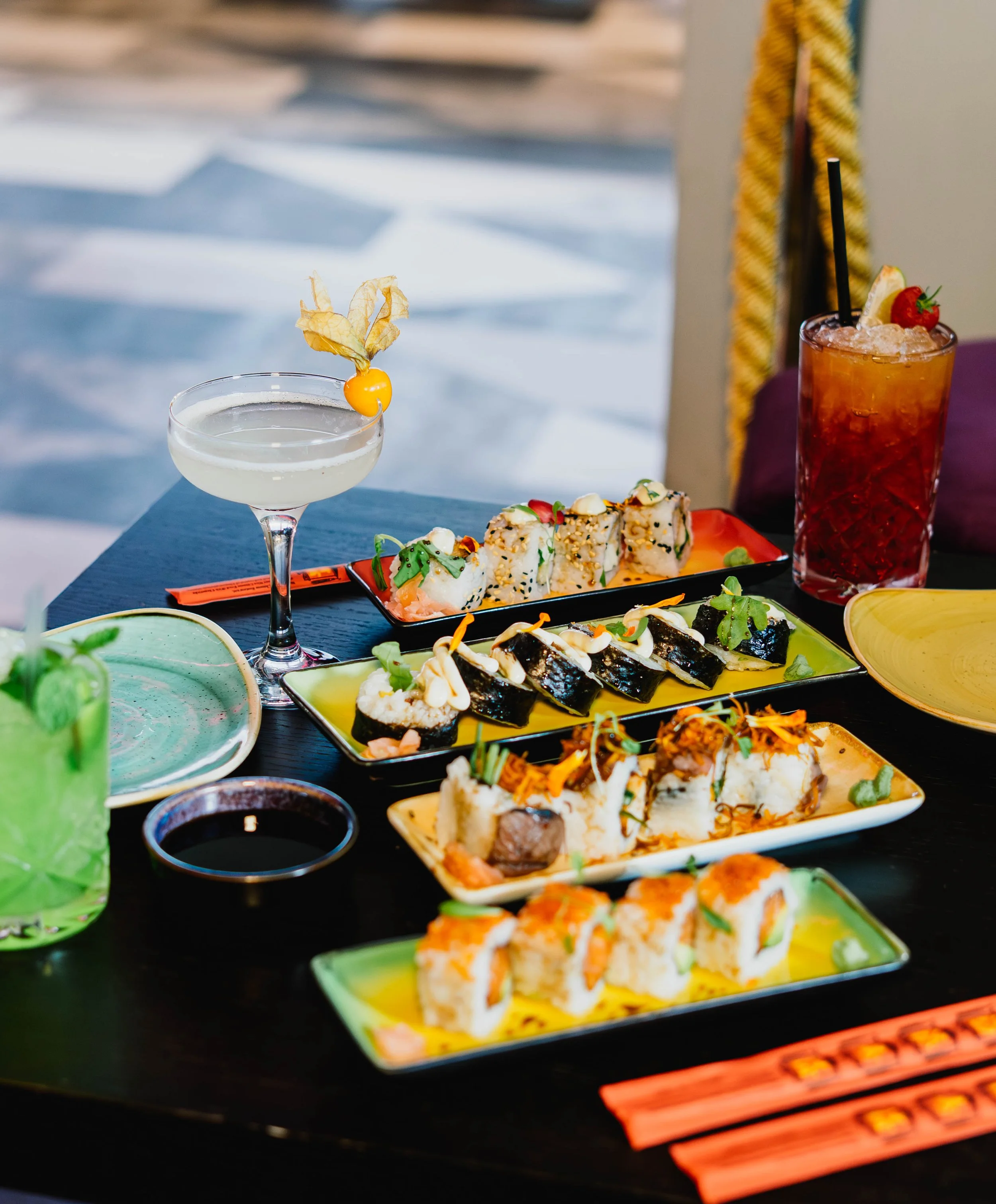 Various plates of sushi, a cocktail with a fruit garnish, a glass of green drink, and a tall glass of strawberry lemonade with a straw and lemon wedge, on a dark table.