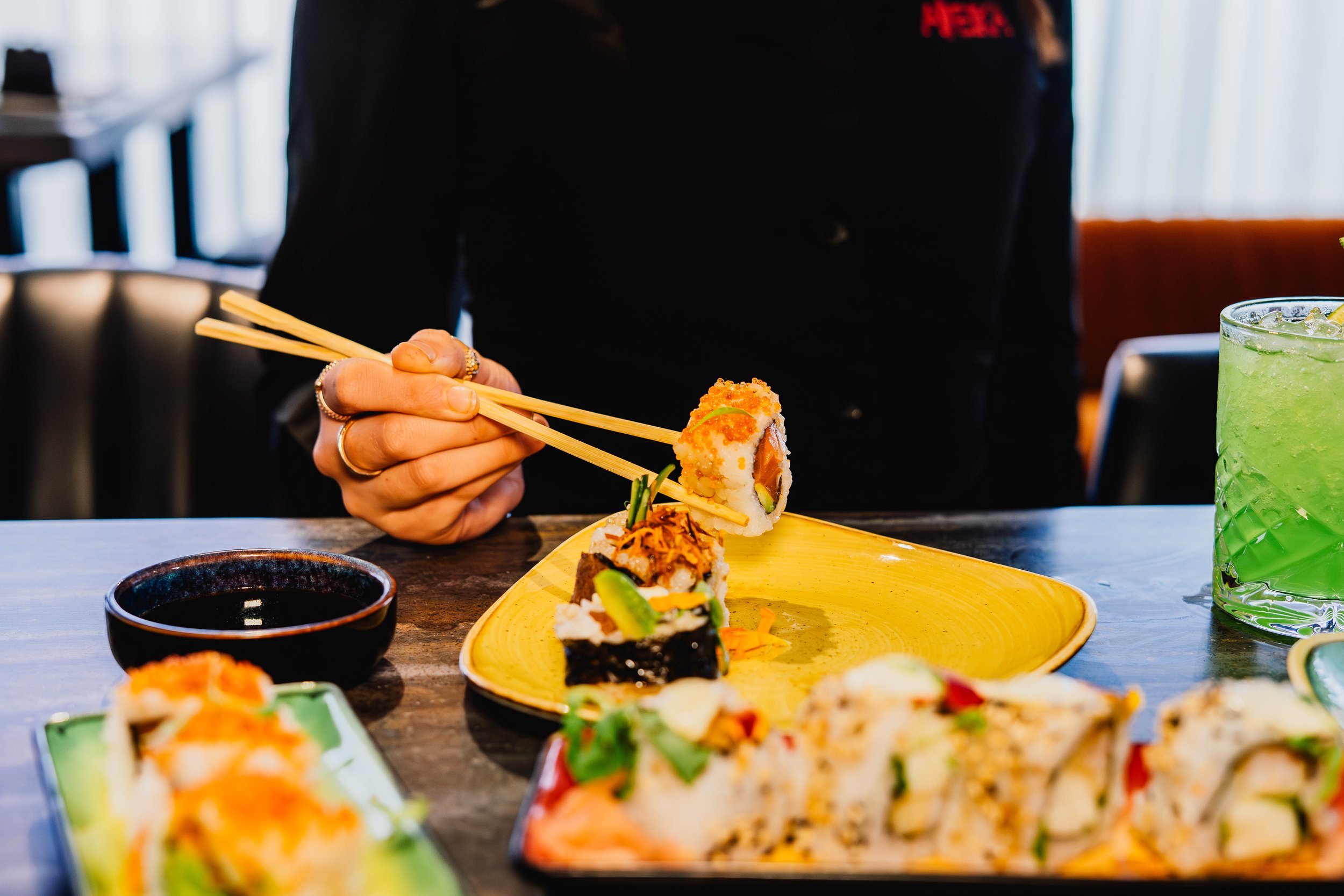 A person in a black jacket holding a pair of chopsticks with sushi on them at a table with various sushi rolls and drinks. The background shows a window with curtains and indoor seating.