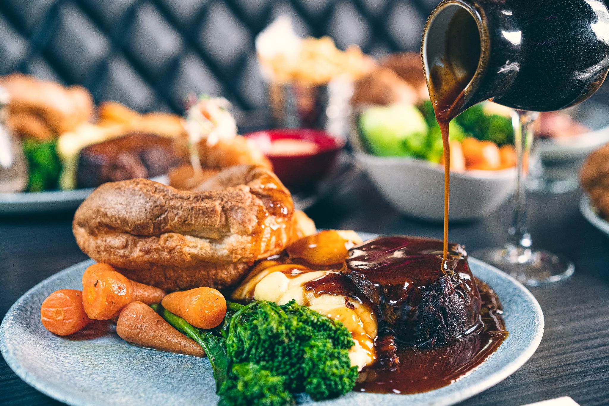 A plate of food with roasted carrots, broccoli, and mashed potatoes topped with gravy, and a slice of meatloaf with gravy. In the background, a close-up of a gravy boat pouring gravy over the meatloaf, with additional dishes on a dark table.