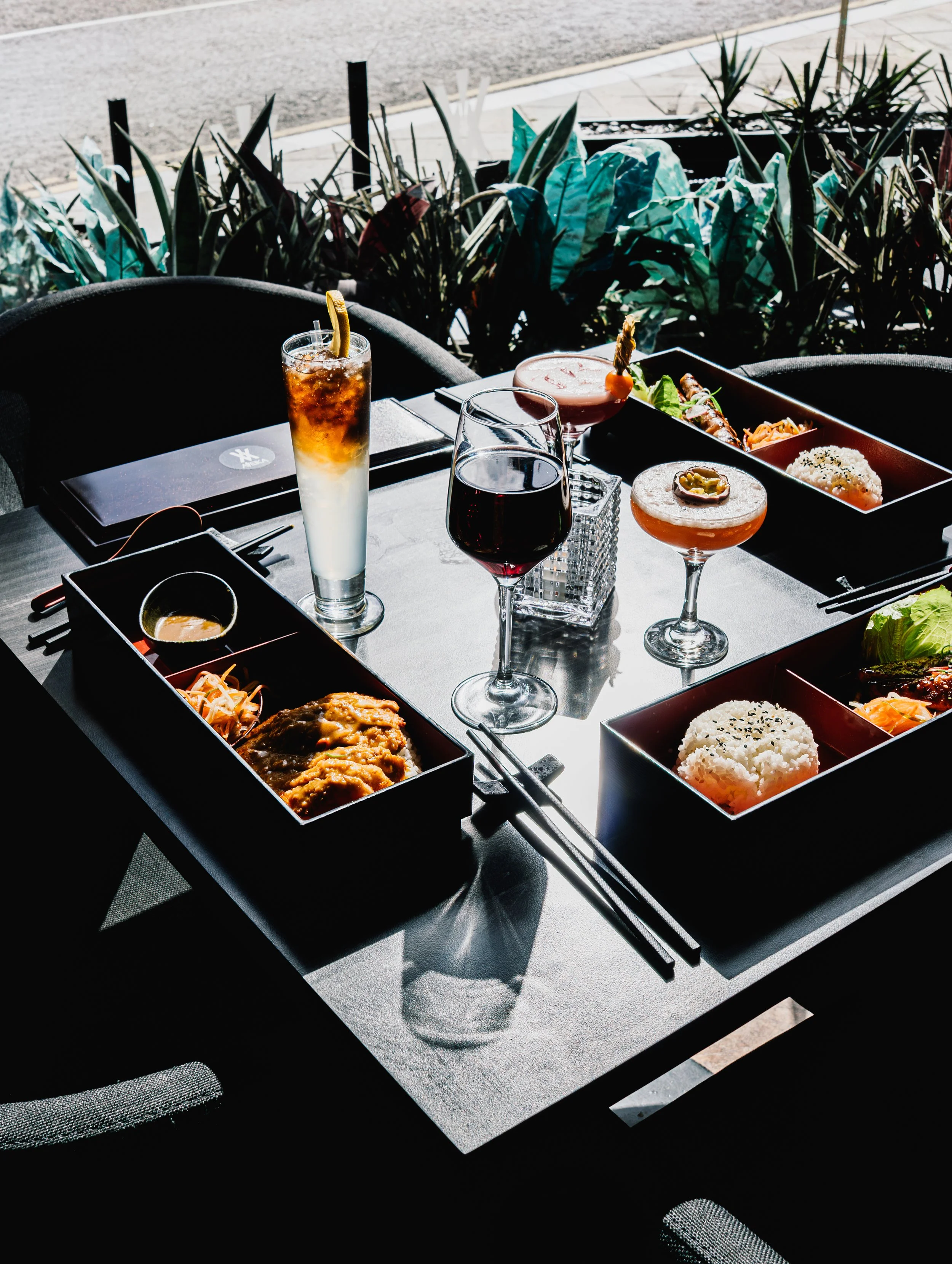Table set with various drinks and food, including cocktails and rice bowls, outdoors with greenery in the background.
